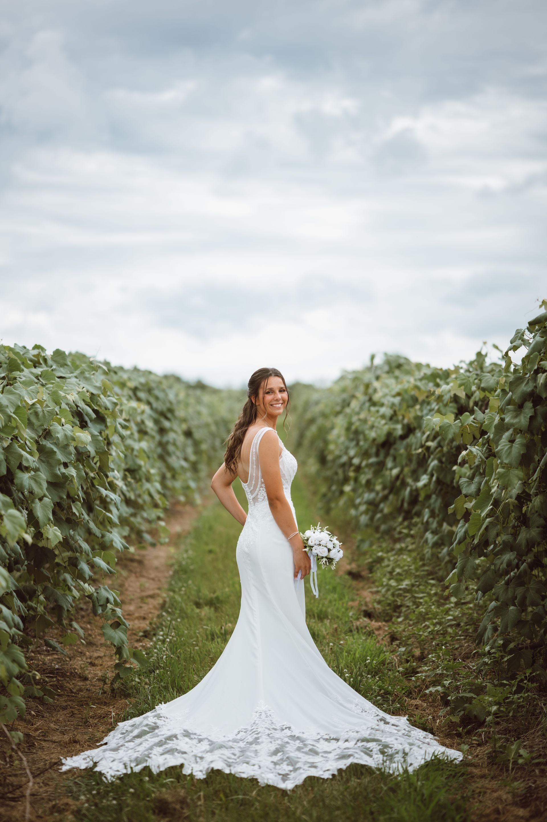 Bride in a white wedding dress smiles, holding bouquet, standing in a vineyard. Cloudy sky.