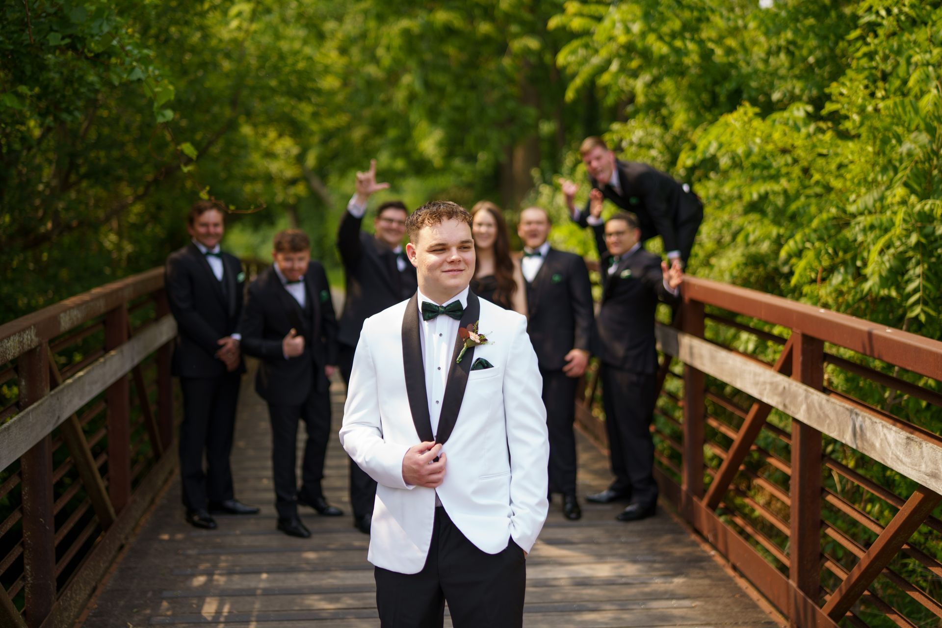 Groom in white tuxedo jacket with groomsmen on bridge in a park, some gesturing exuberantly.