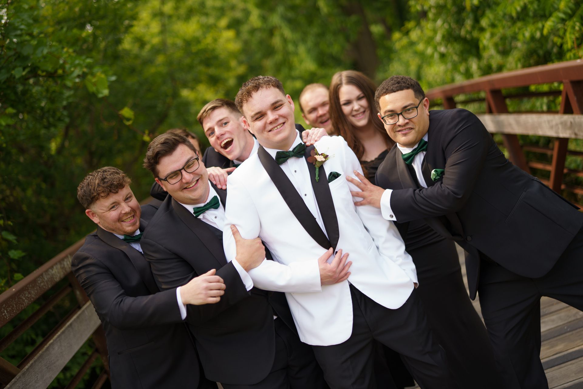 Group of people in formal attire pose playfully on a bridge. Men in suits, groom in white, with excited expressions. Green foliage in background.
