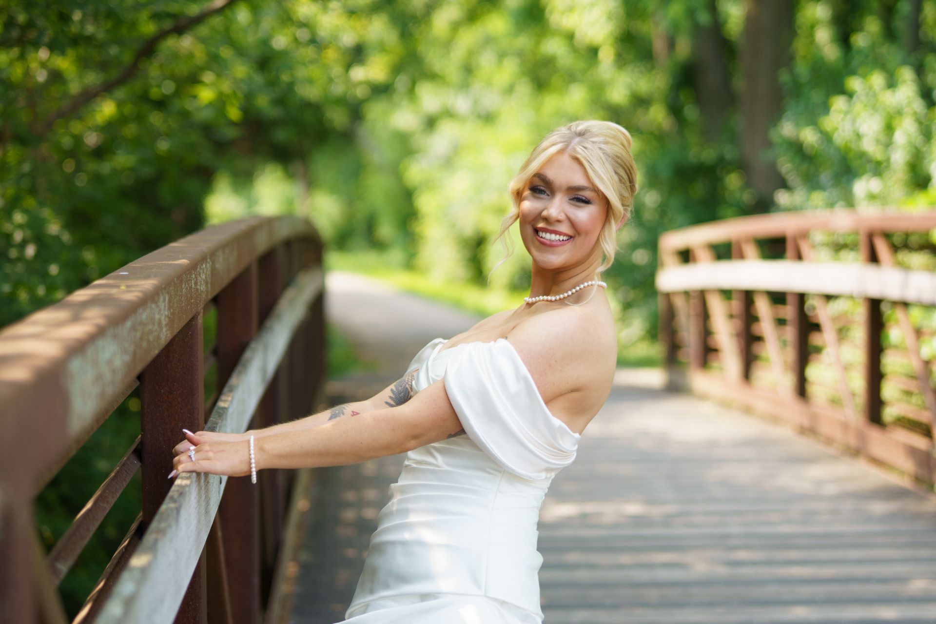 Bride in white dress smiles on a metal bridge in a park.