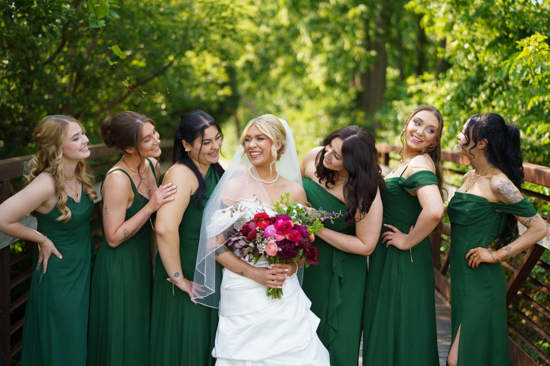 Bride in white dress with bridesmaids in green dresses, smiling on a bridge, holding bouquets.