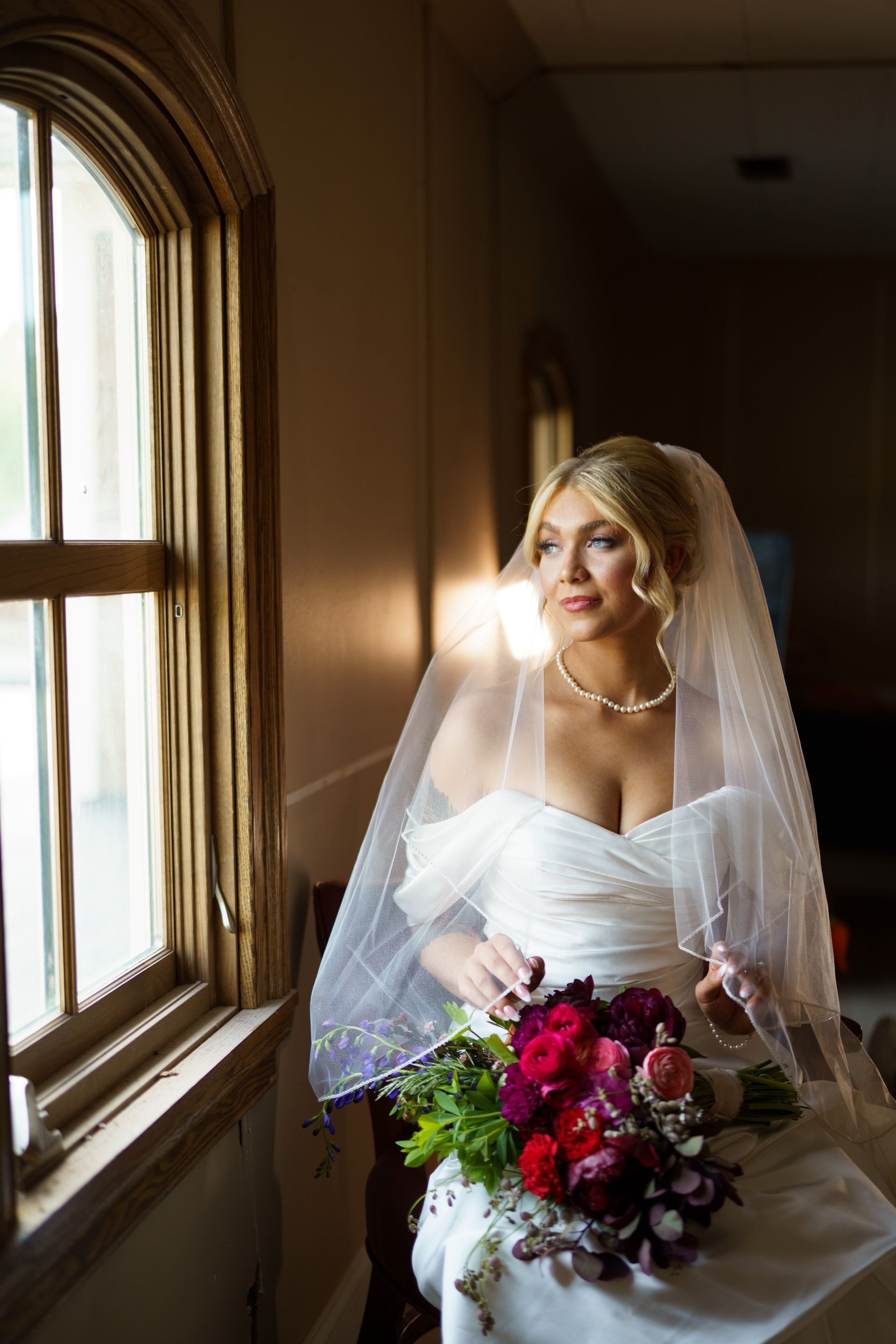 Bride in a wedding dress looking out a window, holding a bouquet, wearing a veil.