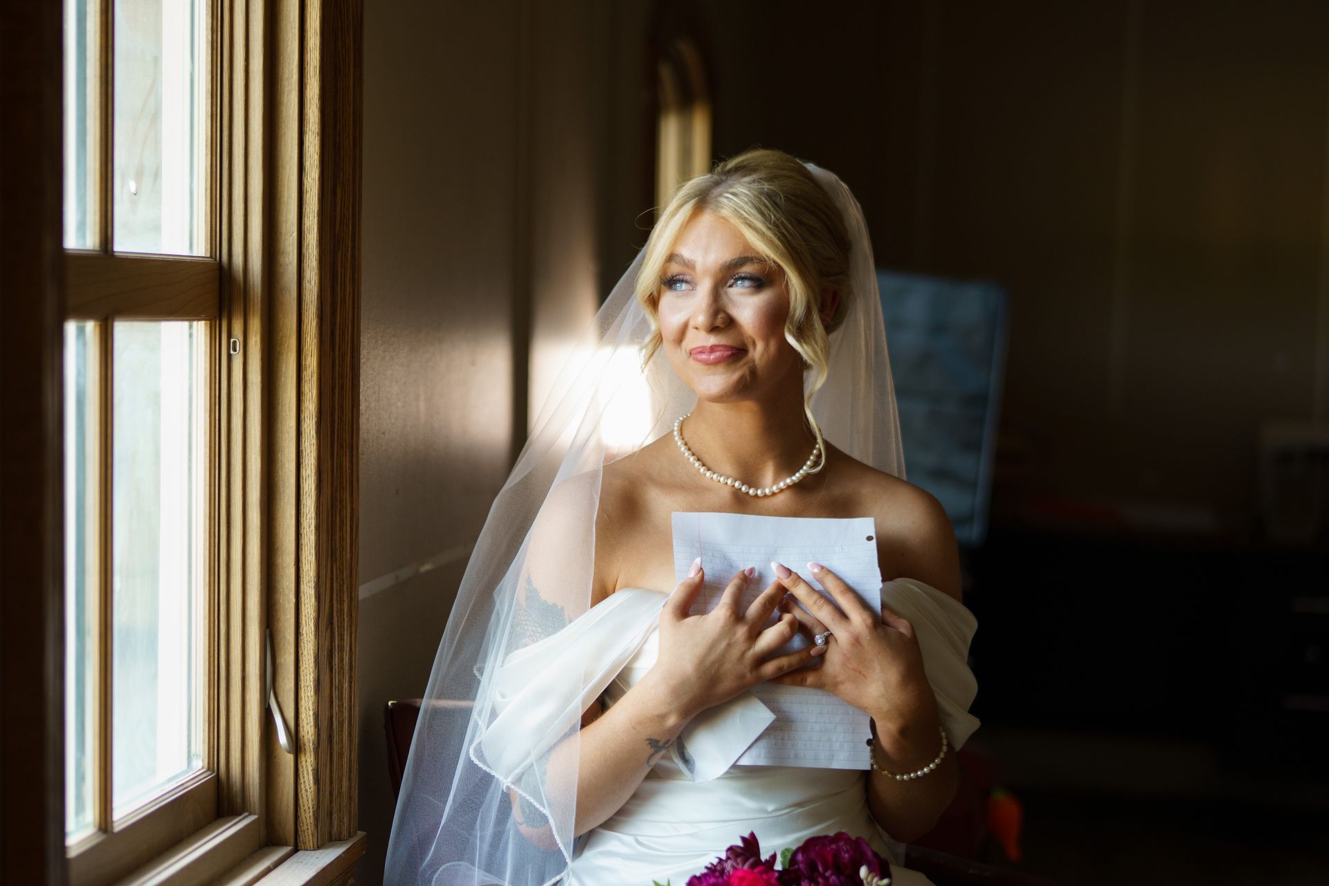 Bride in white gown, holding a letter, looking out a window. Sunlight illuminates her face, pearl necklace visible.