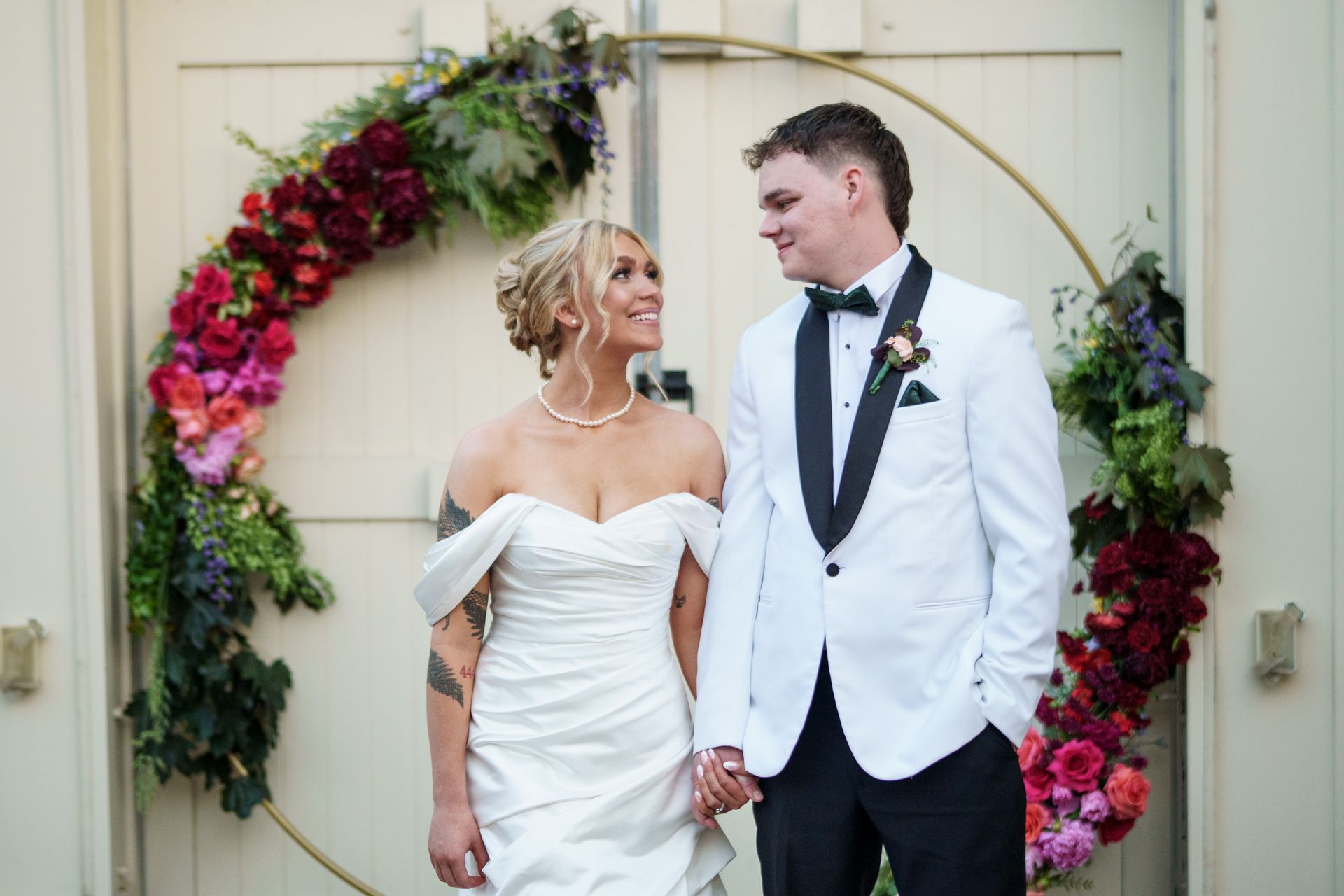 Bride and groom holding hands, smiling at each other. They stand before a floral hoop arch.