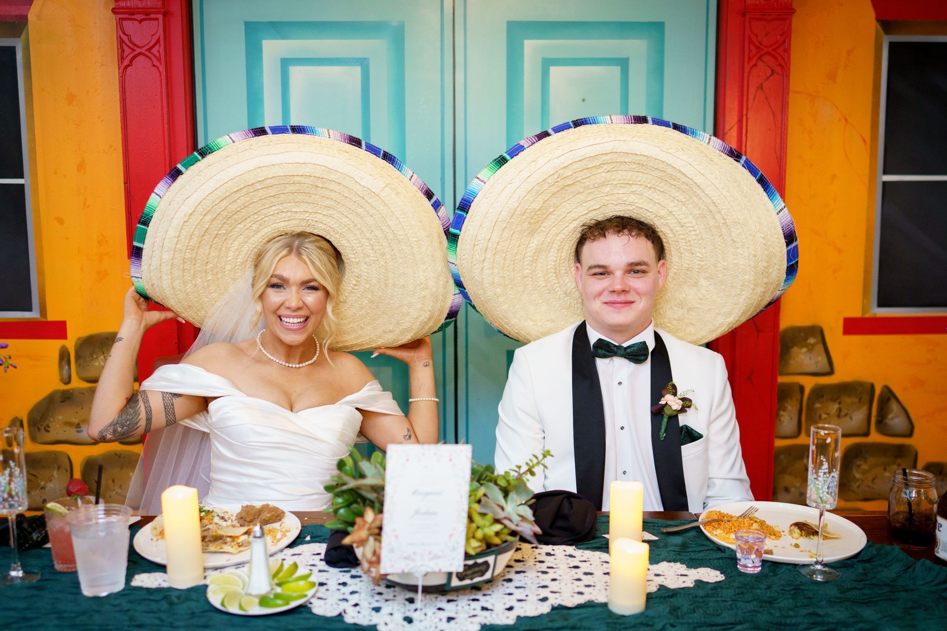 Bride and groom wearing sombreros at a reception table, smiling. Colorful backdrop.