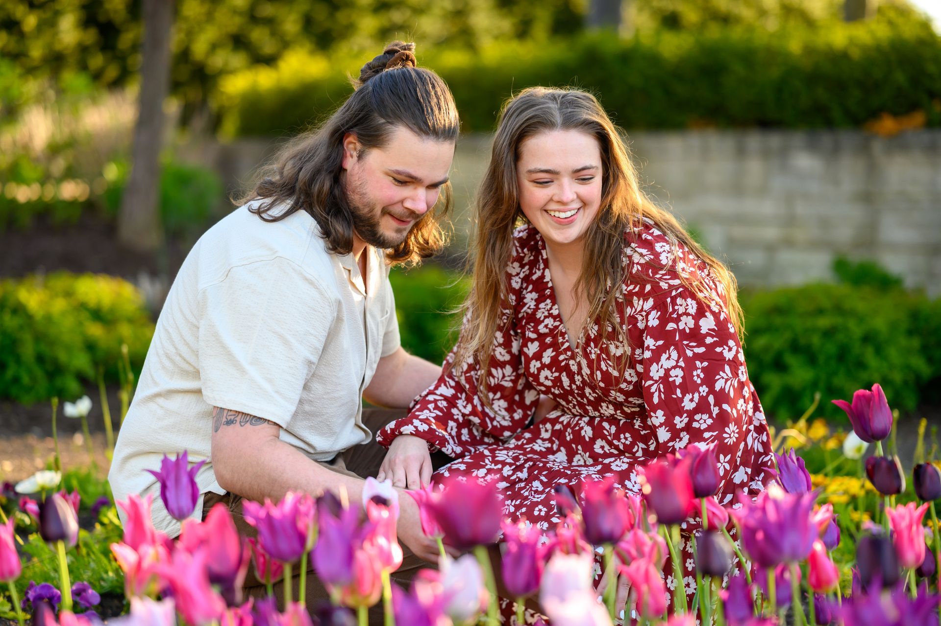 Couple admiring flowers in a garden; woman smiles in a floral dress, man kneels in cream shirt; purple and pink tulips.