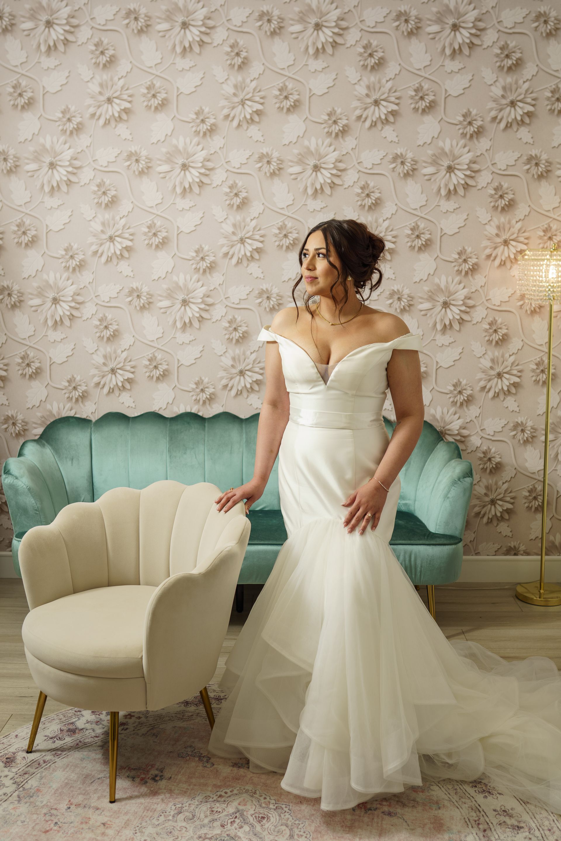Woman in a white wedding dress poses near a teal sofa and cream chair. She looks to the side in a studio.
