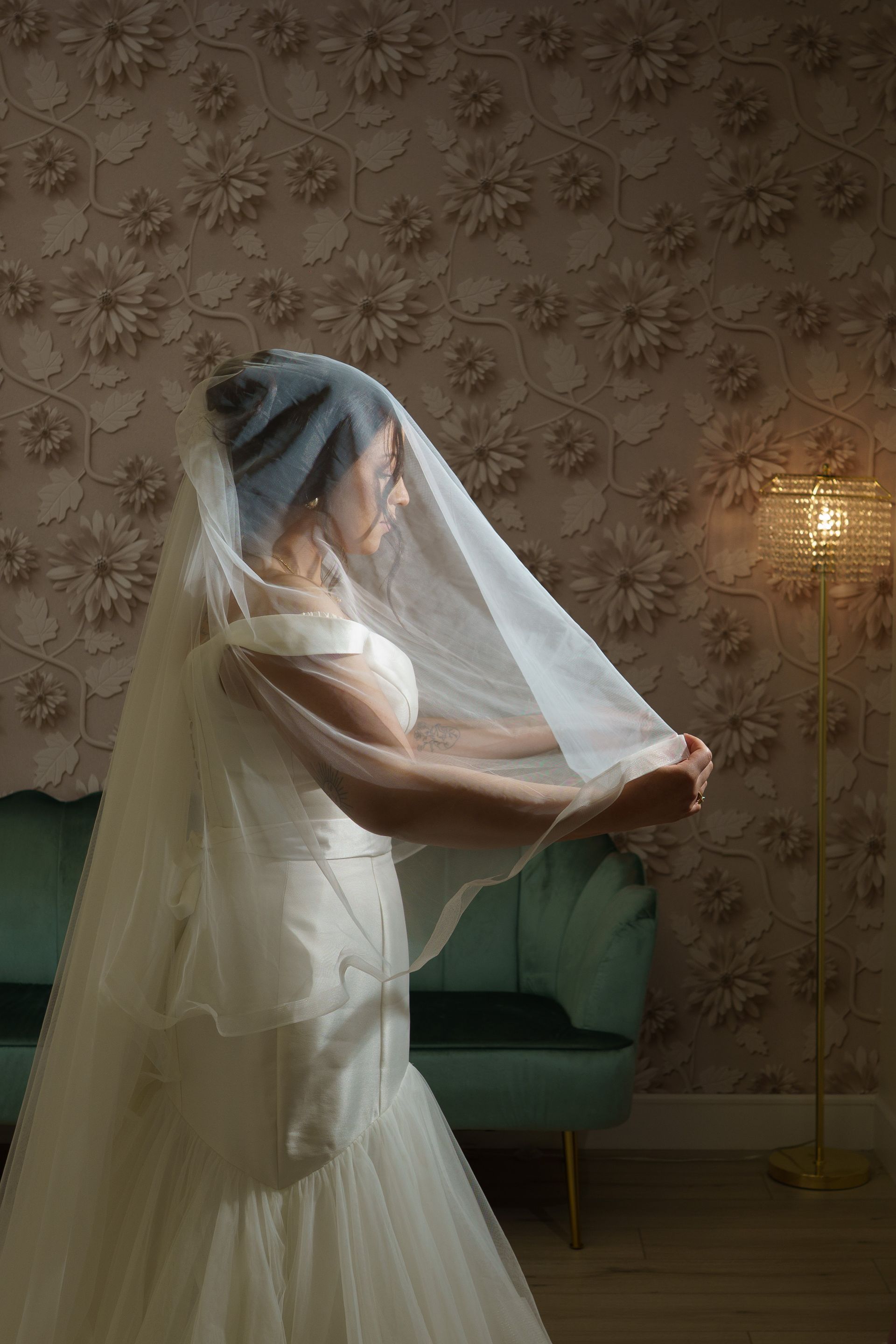 Bride in a wedding dress, lifting a sheer veil, stands near a teal sofa in a room with floral wallpaper and a floor lamp.
