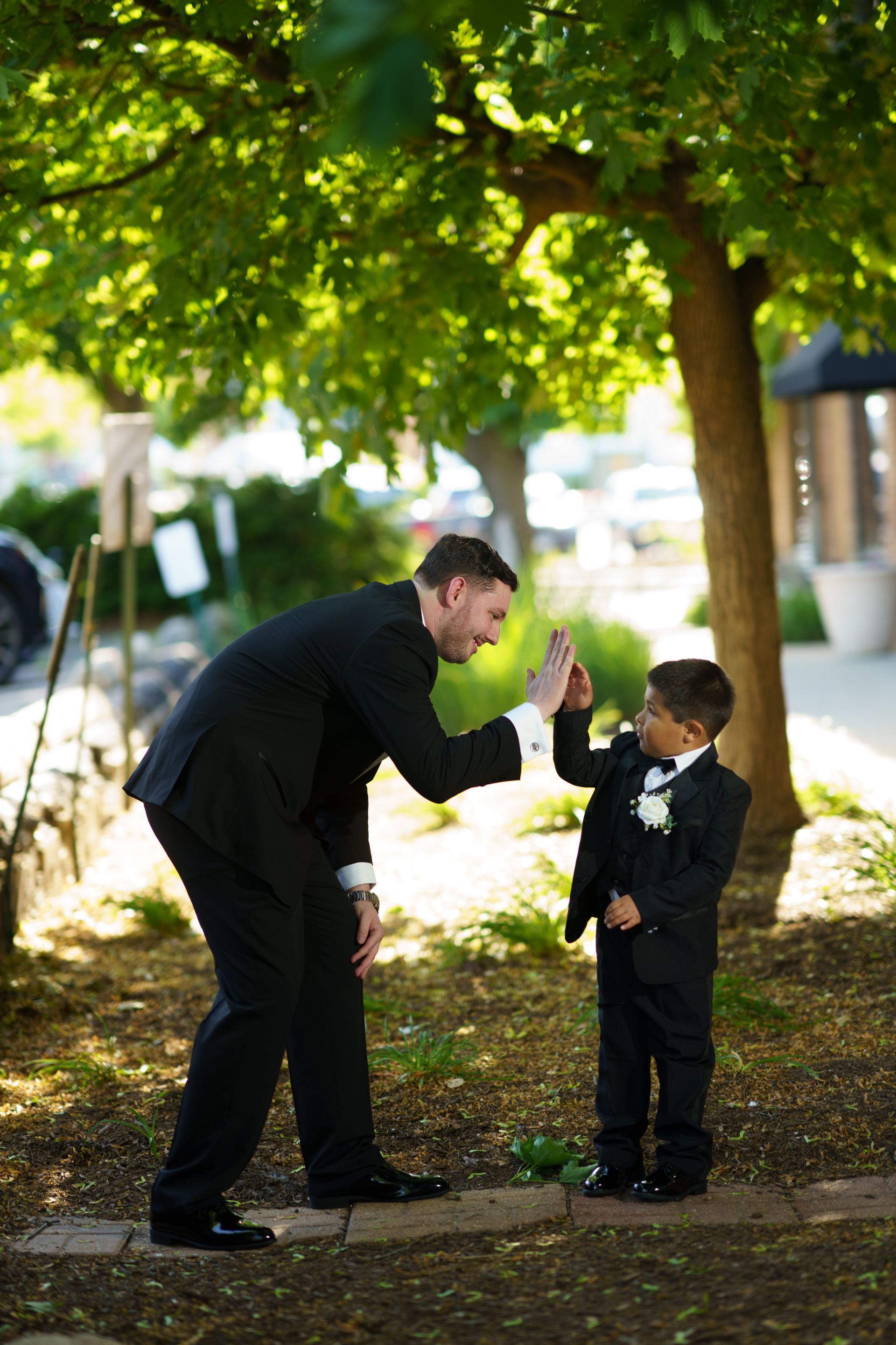 Man in suit high-fives a boy in a suit outdoors under a tree, both smiling.