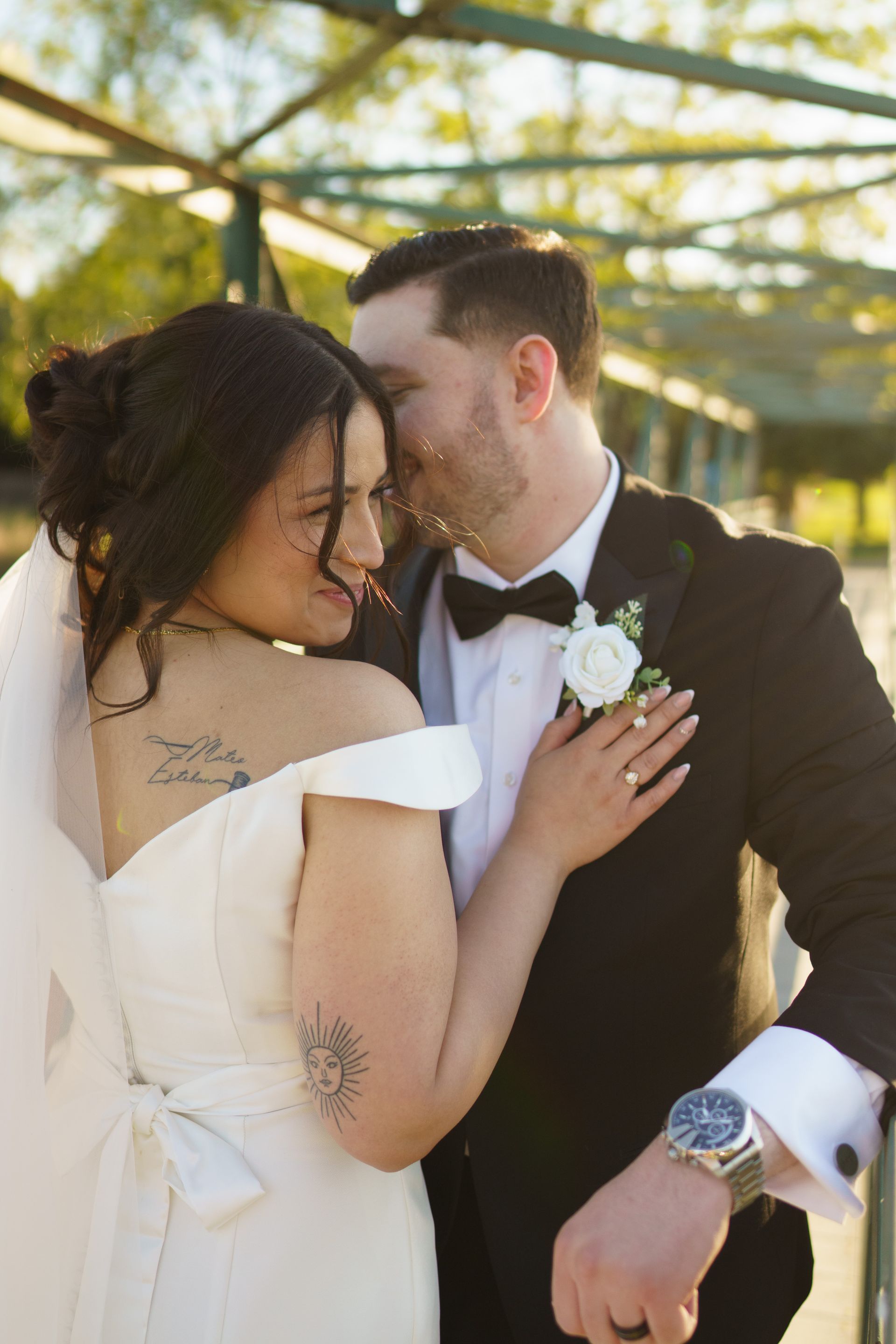 Bride and groom embrace outdoors, she wears white dress and veil, he wears a tuxedo, both smiling.