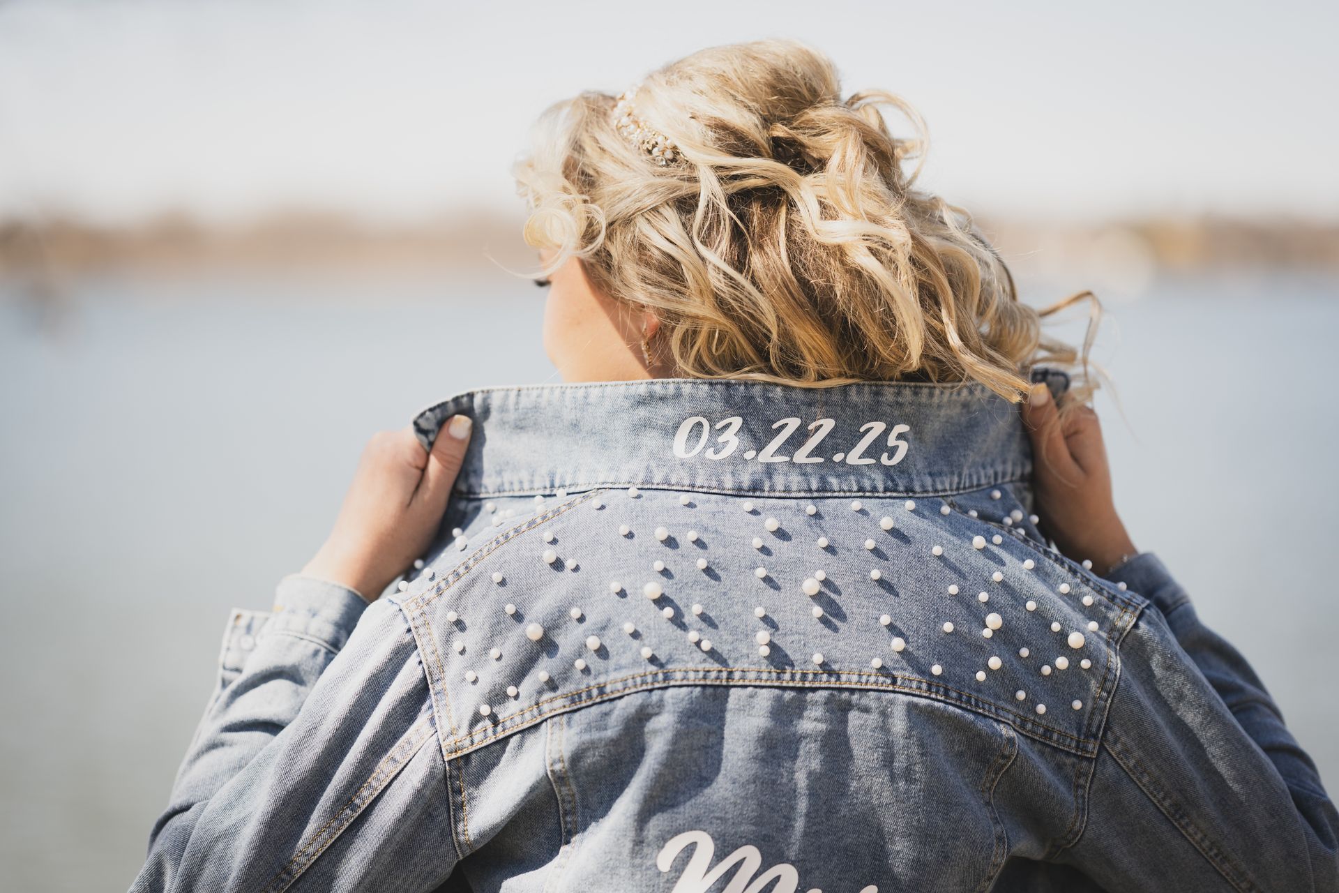 Woman wearing a denim jacket with pearls and wedding date 03.22.25 embroidered on it, near water.