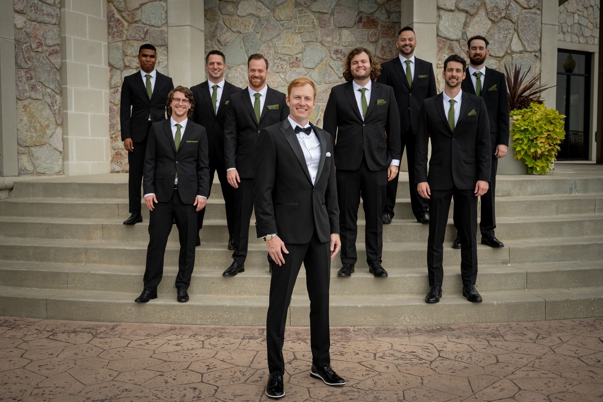 Group of men in black suits with green accents posing on steps outside.
