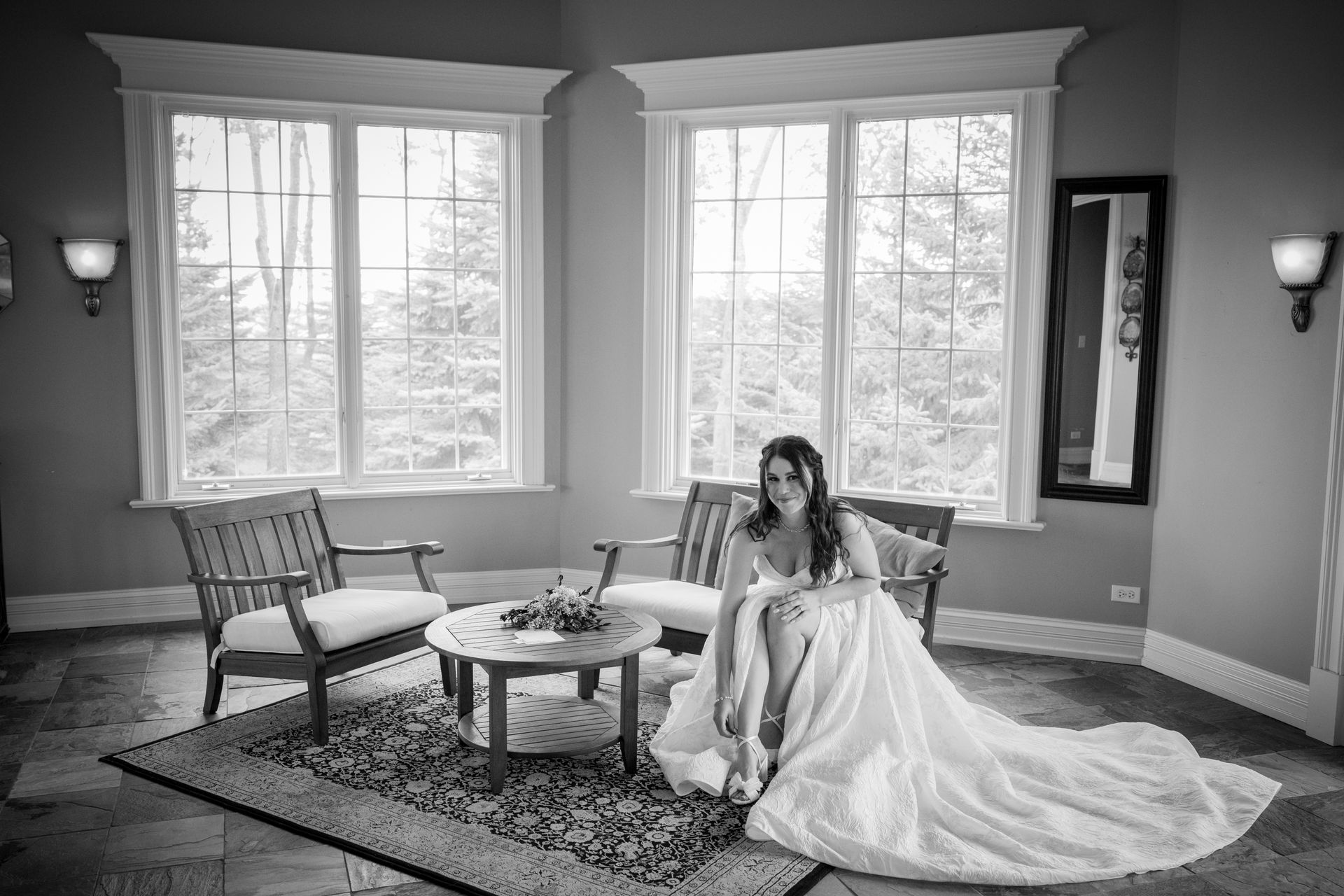 Bride in a wedding dress sits by a table in a room with large windows, smiling.