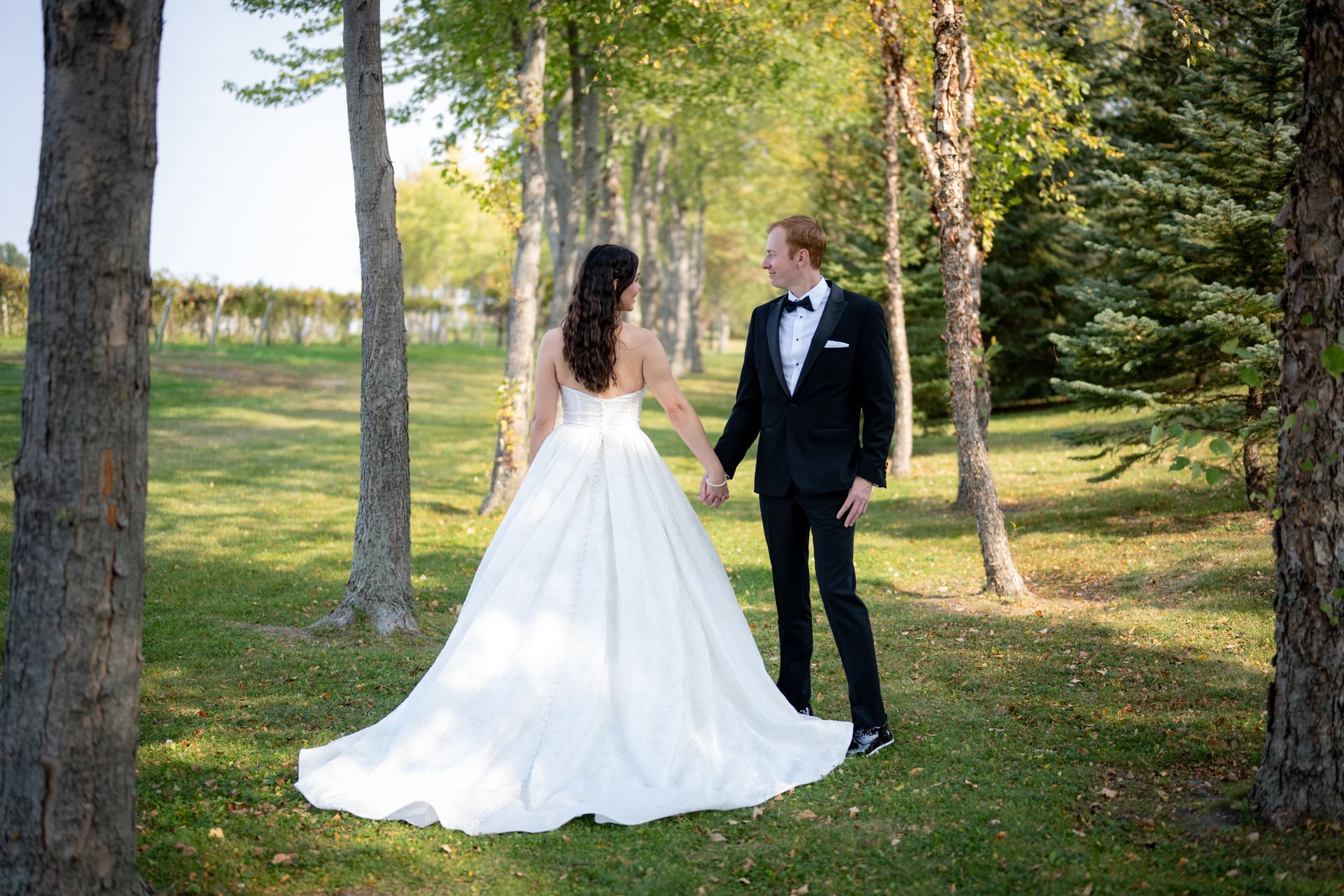 Bride and groom holding hands, standing in a tree-lined grassy area, facing away from the camera.