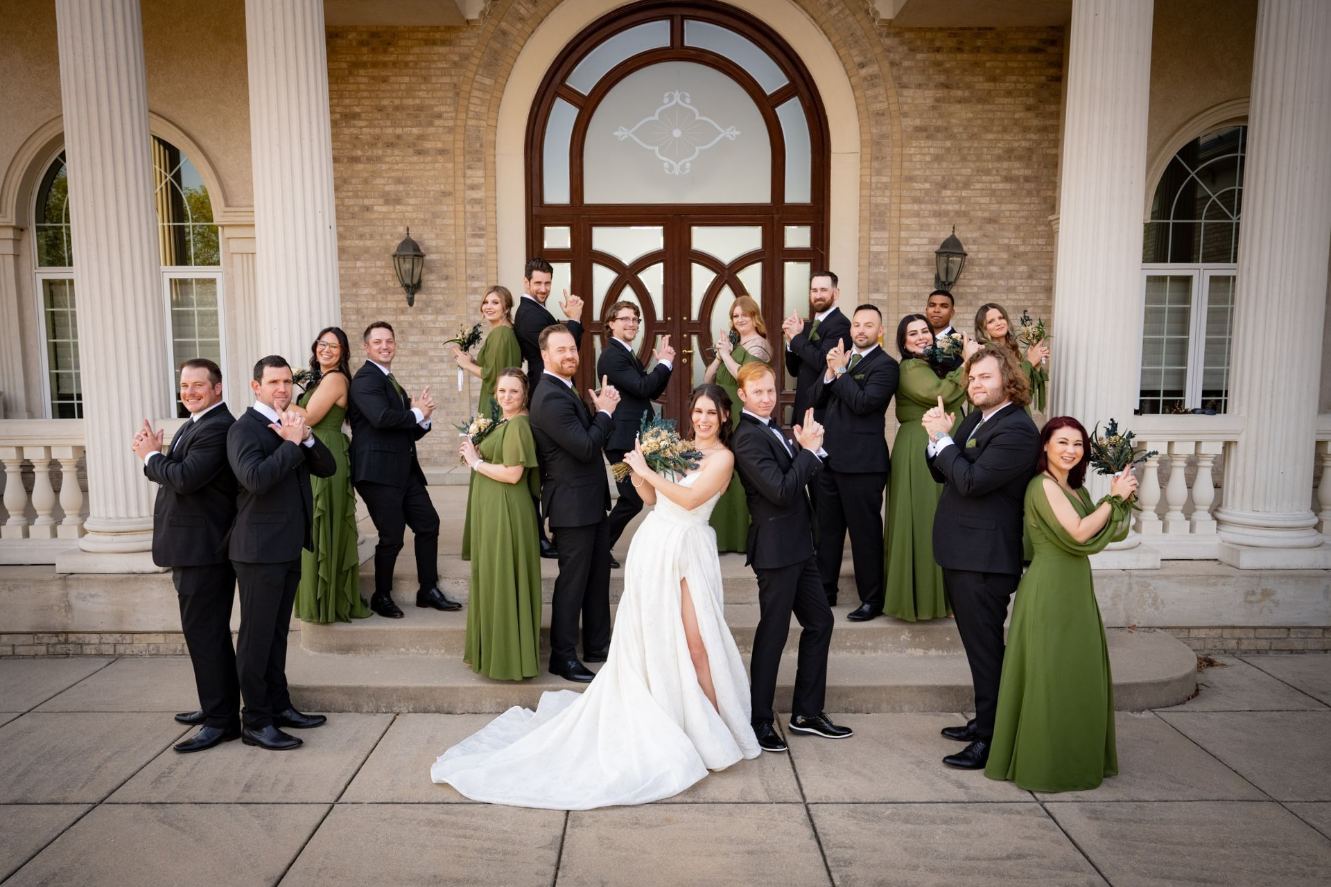 Wedding party posing on steps of a building. Bride in white dress, bridesmaids in green, groomsmen in black, applauding.