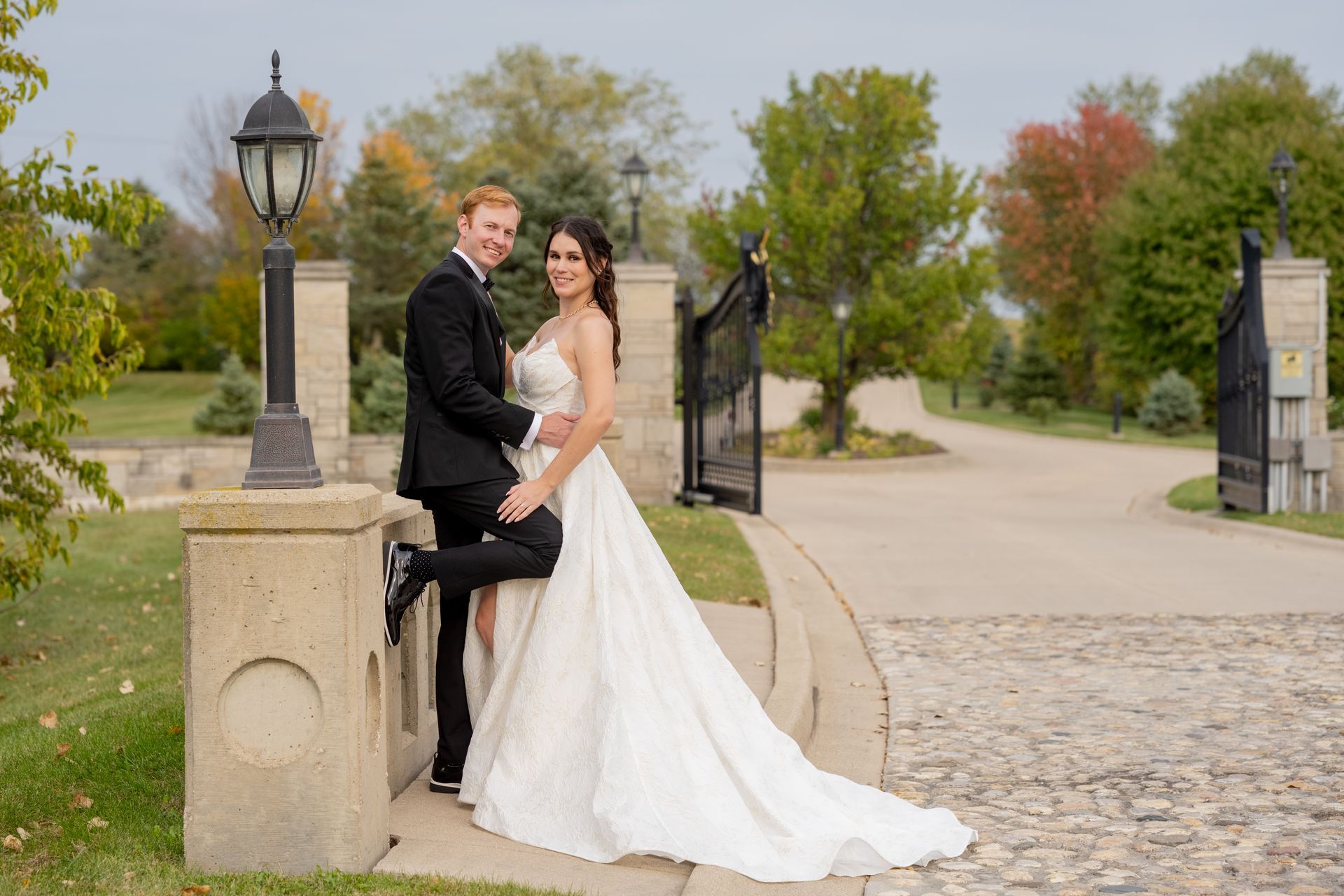 Couple in wedding attire poses by stone structure near wrought iron gate.