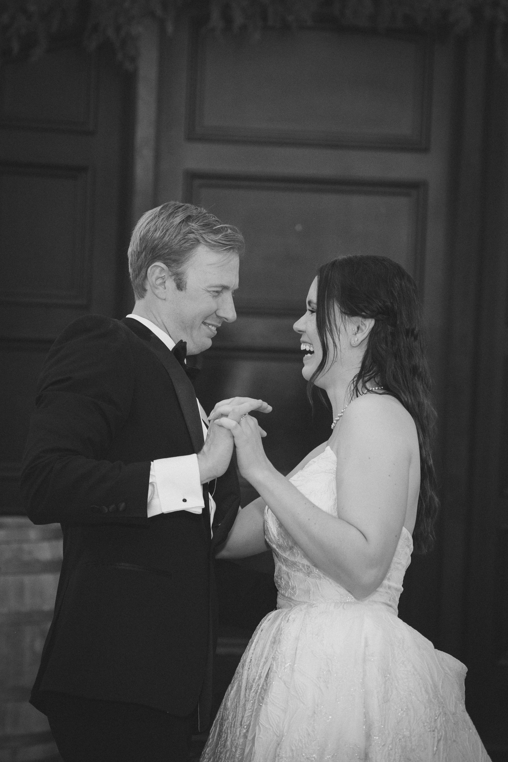 Bride and groom laughing while holding hands during first dance; tuxedo, wedding dress.