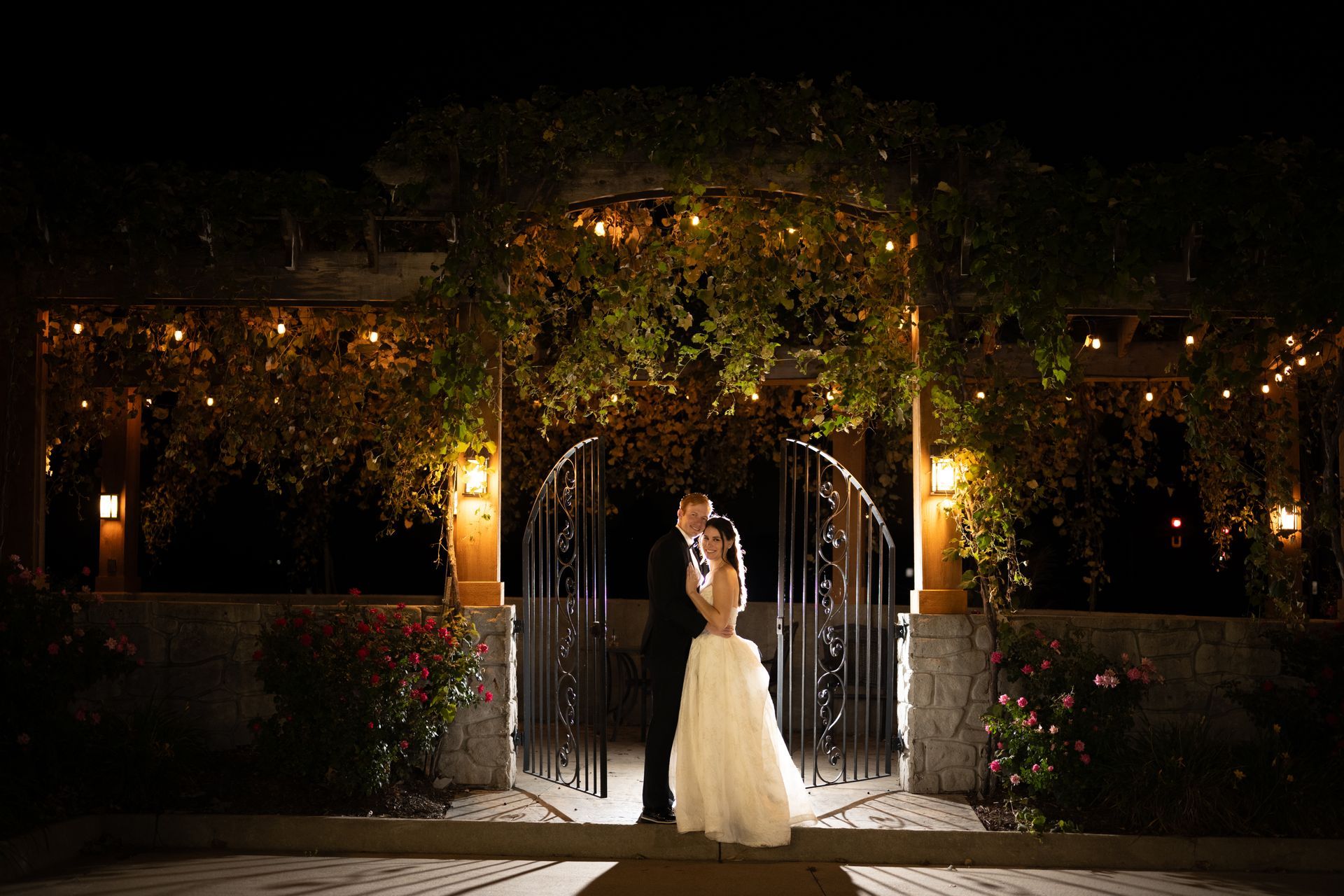 Couple kissing under an archway adorned with lights and greenery, at night.