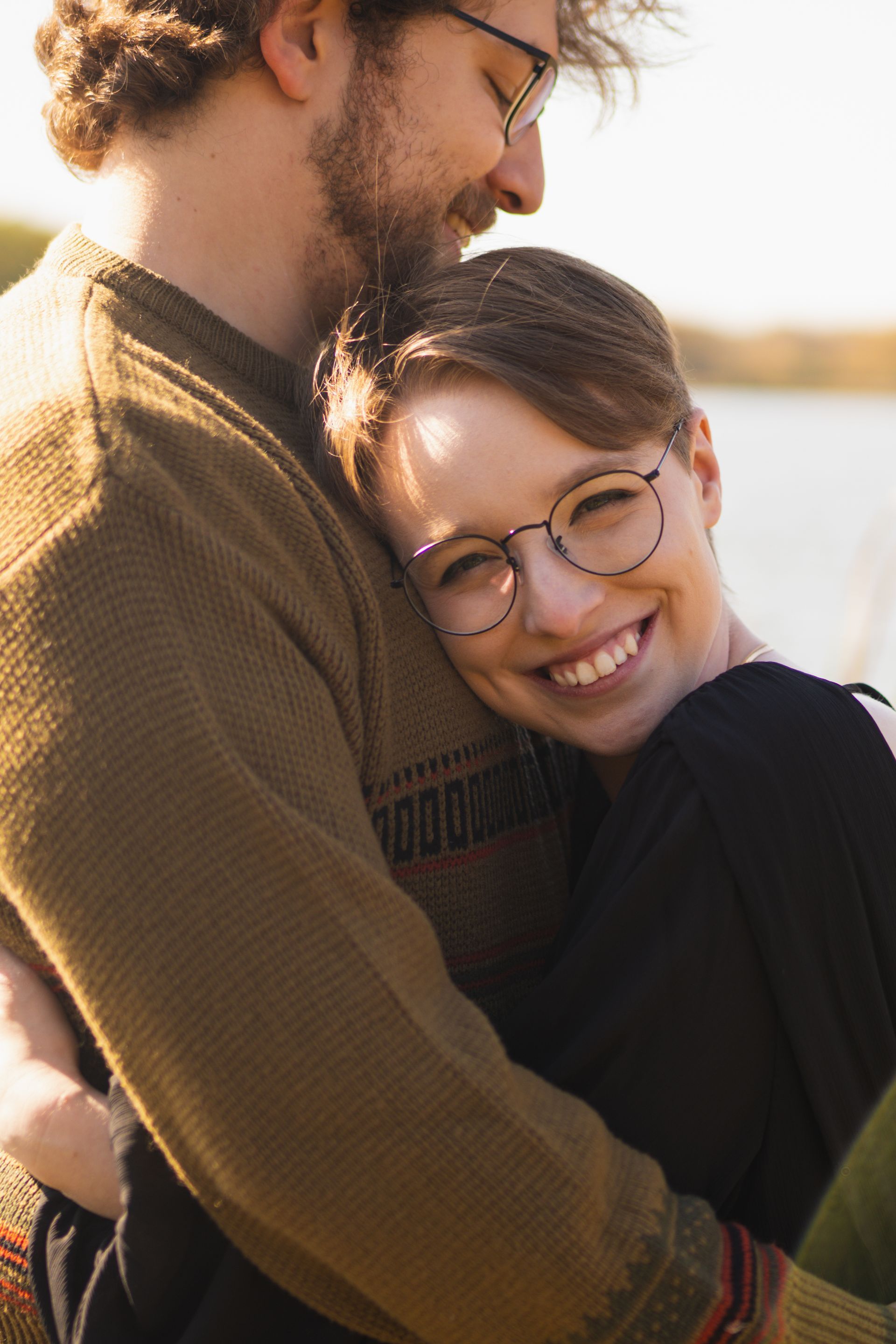 Man hugs woman; both wear glasses. Woman smiles, head on his shoulder, outdoors near water.
