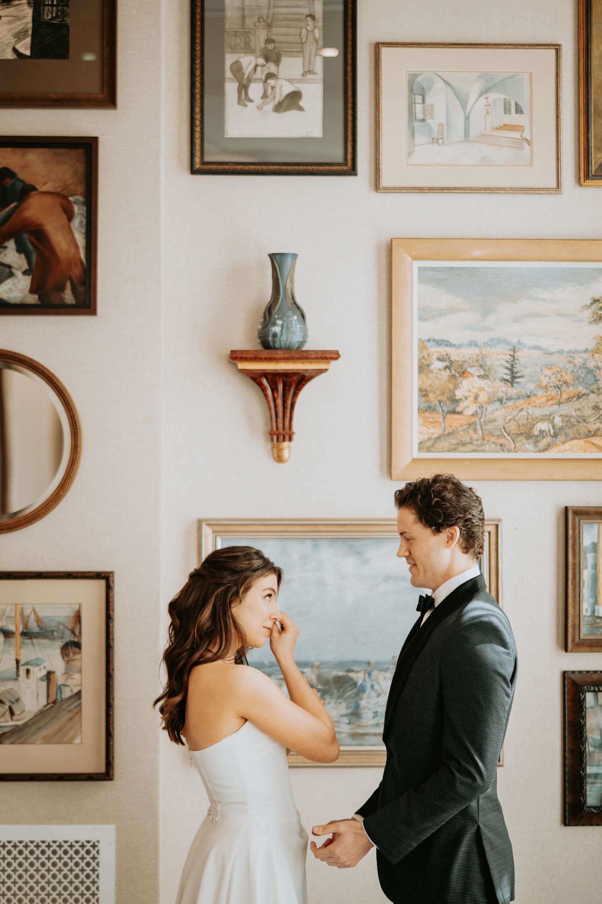 Bride and groom facing each other, looking at each other, in front of a wall of framed art.