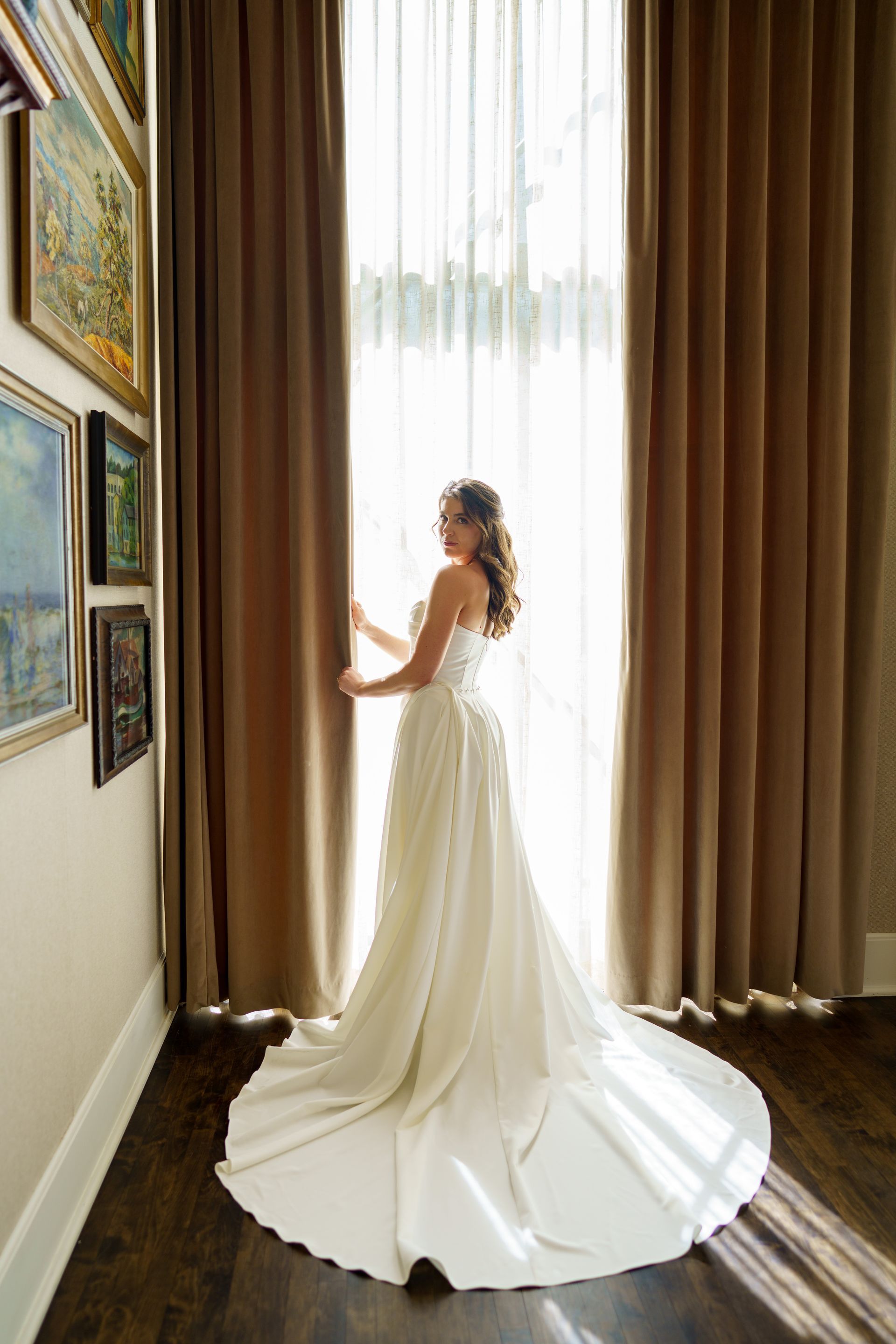 Bride in a white gown standing near a window, holding the curtain. A wall of art is on her left.
