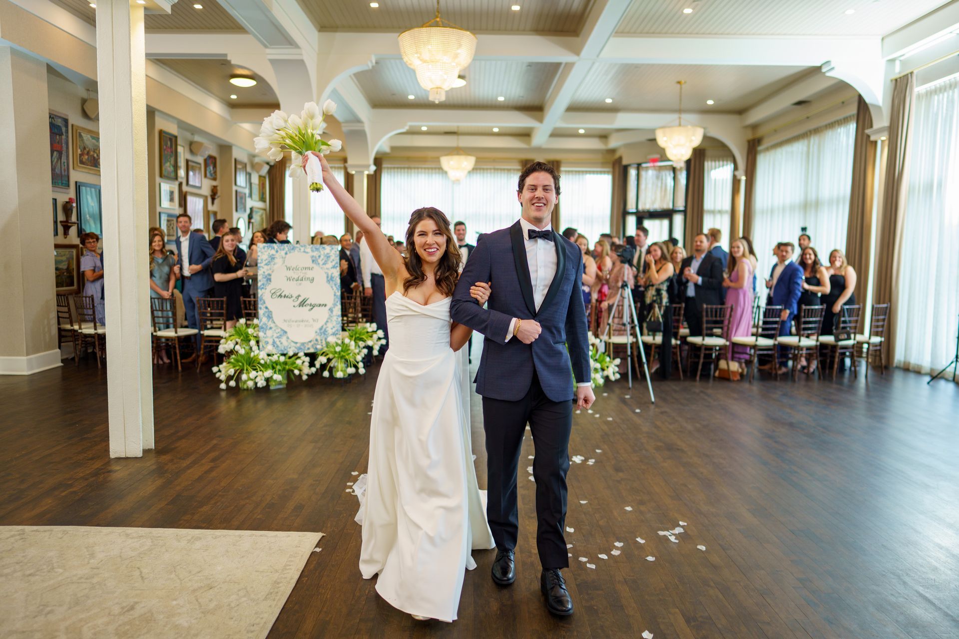 Bride and groom exiting wedding ceremony, arm in arm, smiling, holding flowers, surrounded by guests.