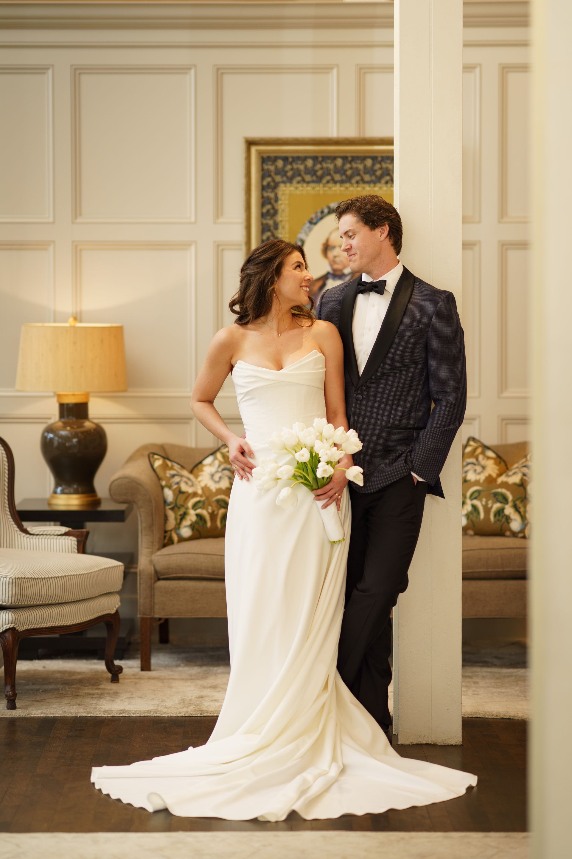 Bride and groom in formal attire, smiling at each other. Bride holds bouquet, groom leans against column in ornate room.