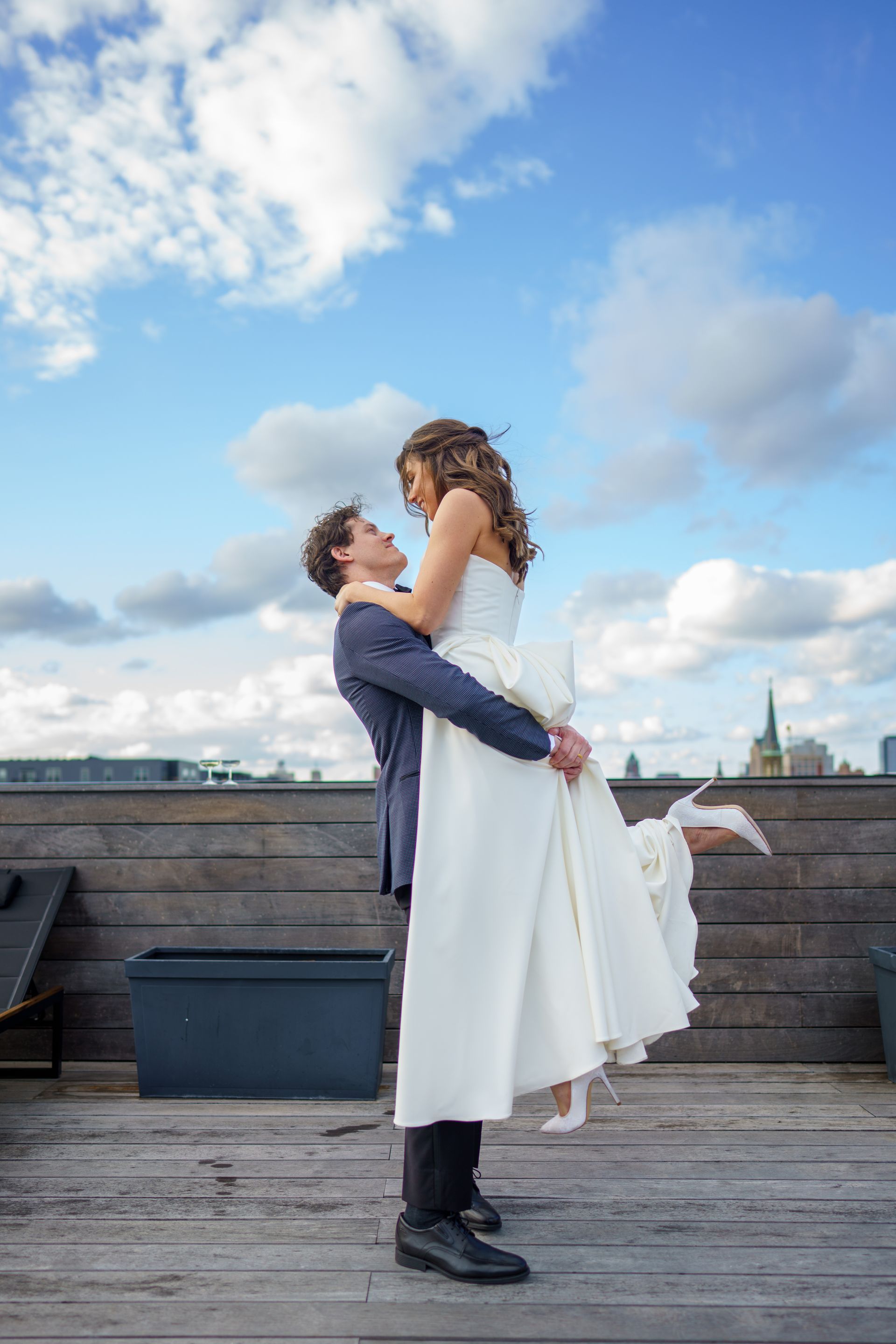 Groom lifts bride on rooftop, blue sky, white dress.