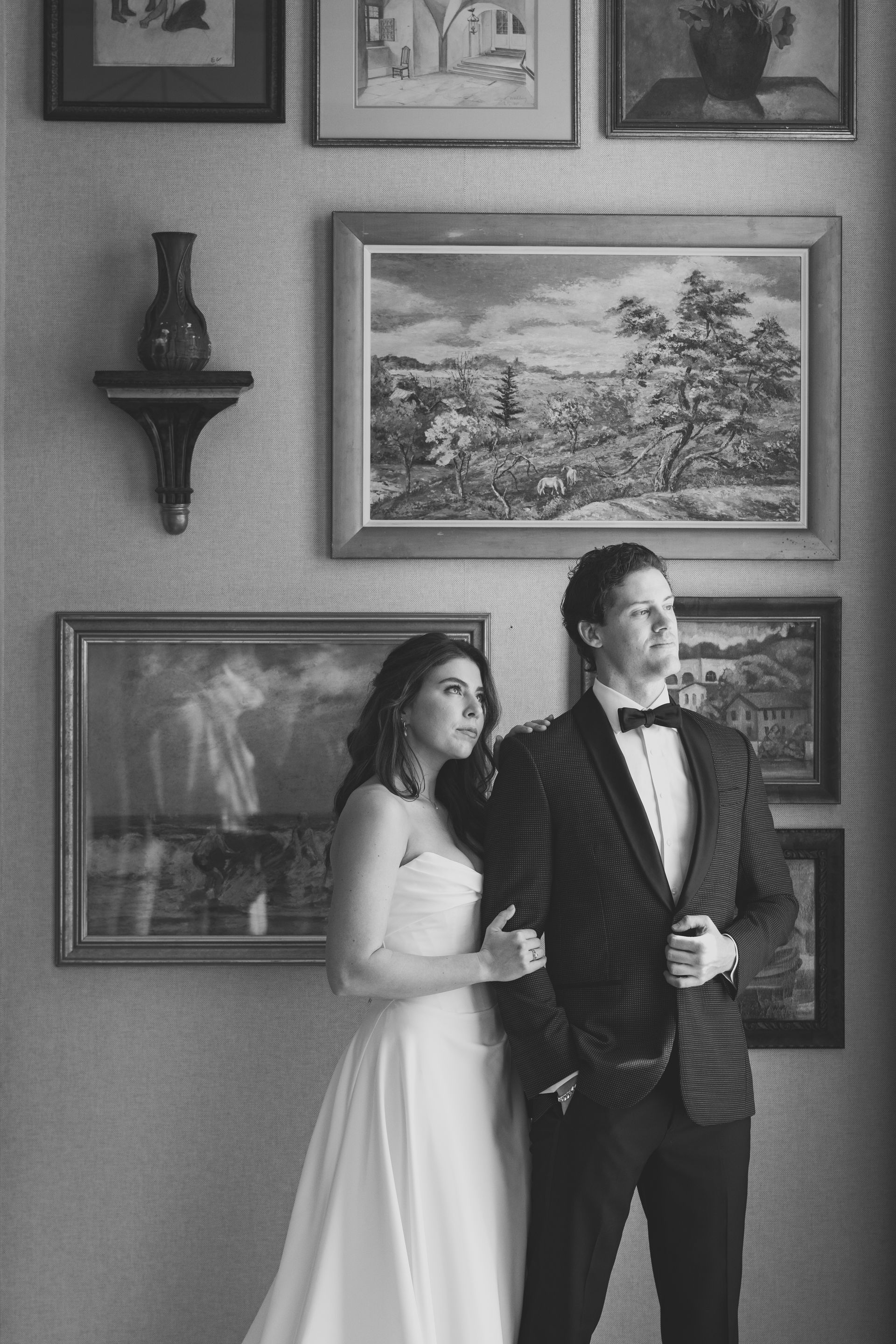 Couple in formal wear poses in front of a wall with artwork. Woman in a white gown; man in a tuxedo.
