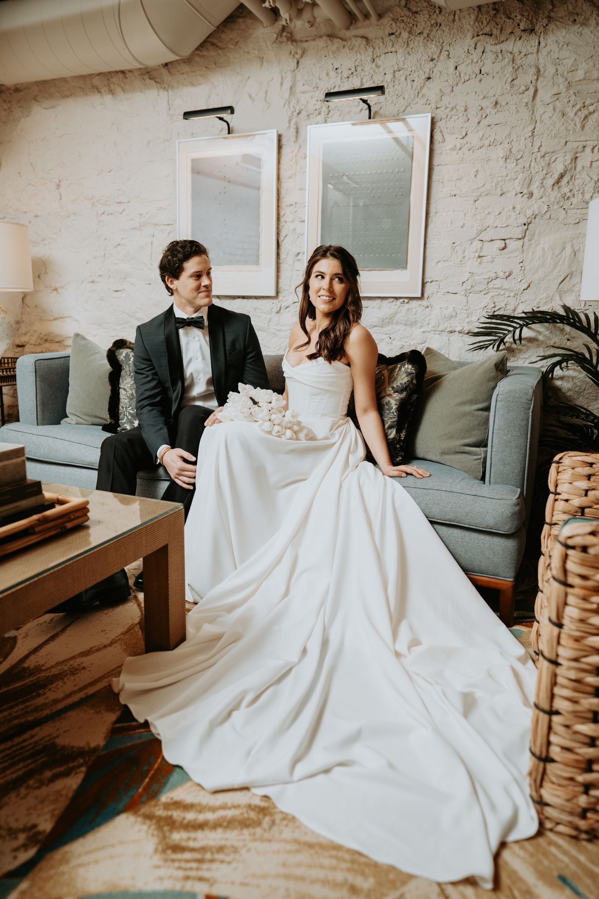 Bride and groom seated on a blue couch, bride in a white gown, groom in a tuxedo, interior setting.