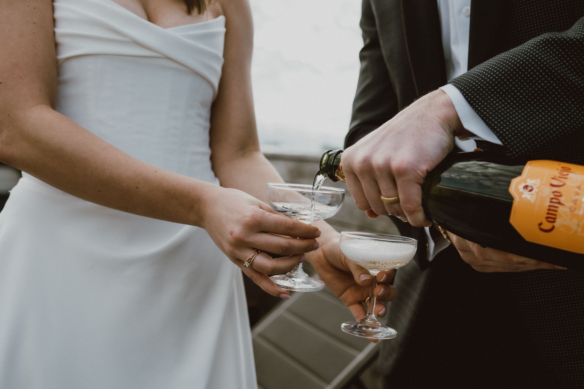Couple pouring champagne into glasses at an outdoor wedding, wearing wedding attire.