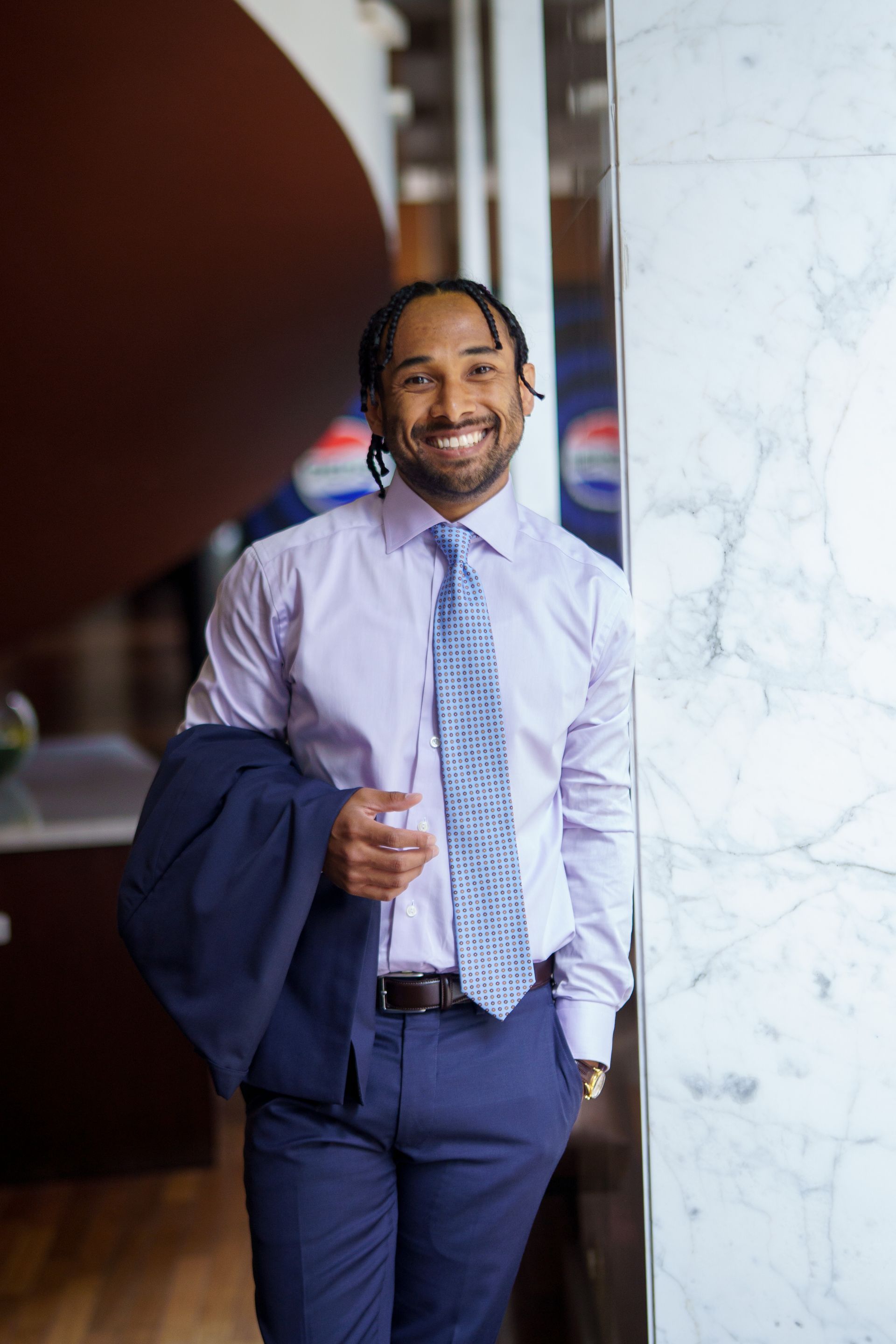 Man in a light purple shirt, blue tie, and suit pants smiling, holding a jacket and glass near a marble pillar.