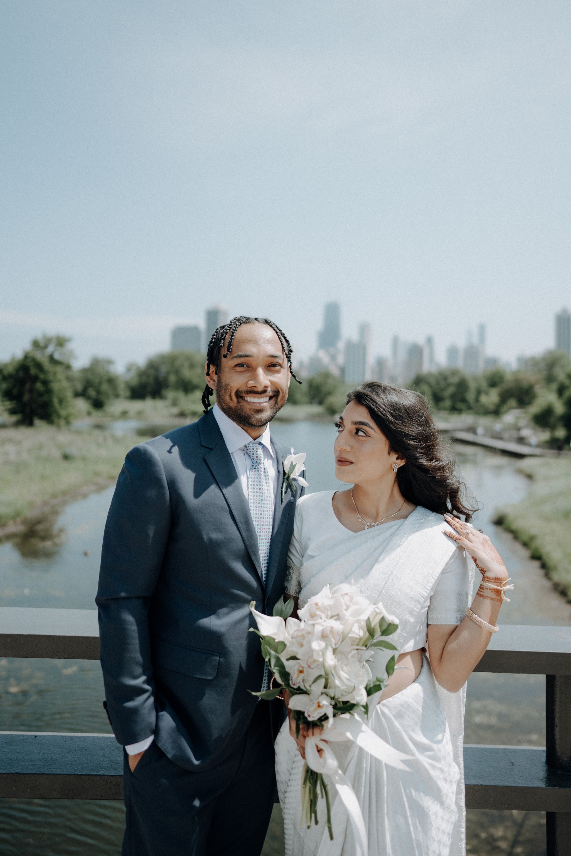 Couple in wedding attire on bridge, city skyline in background; sunny day.