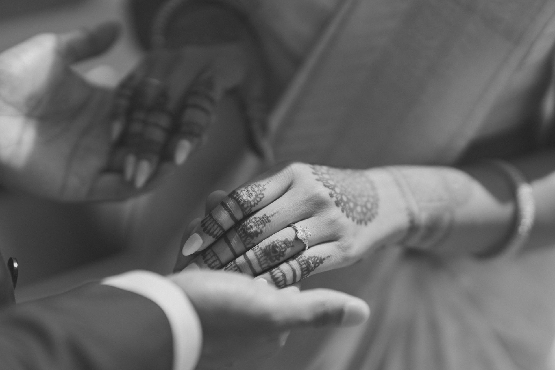 Hands of a couple, one with henna, hold each other's. The scene is in black and white.