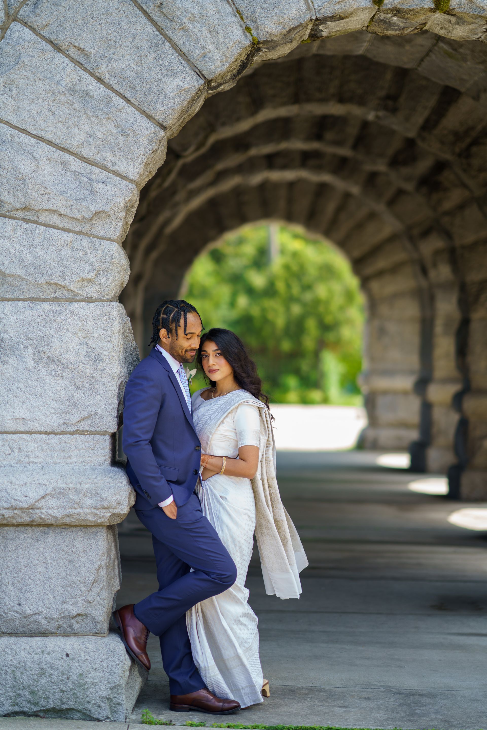 Couple in formal attire pose under a stone archway, woman in white saree, man in blue suit.