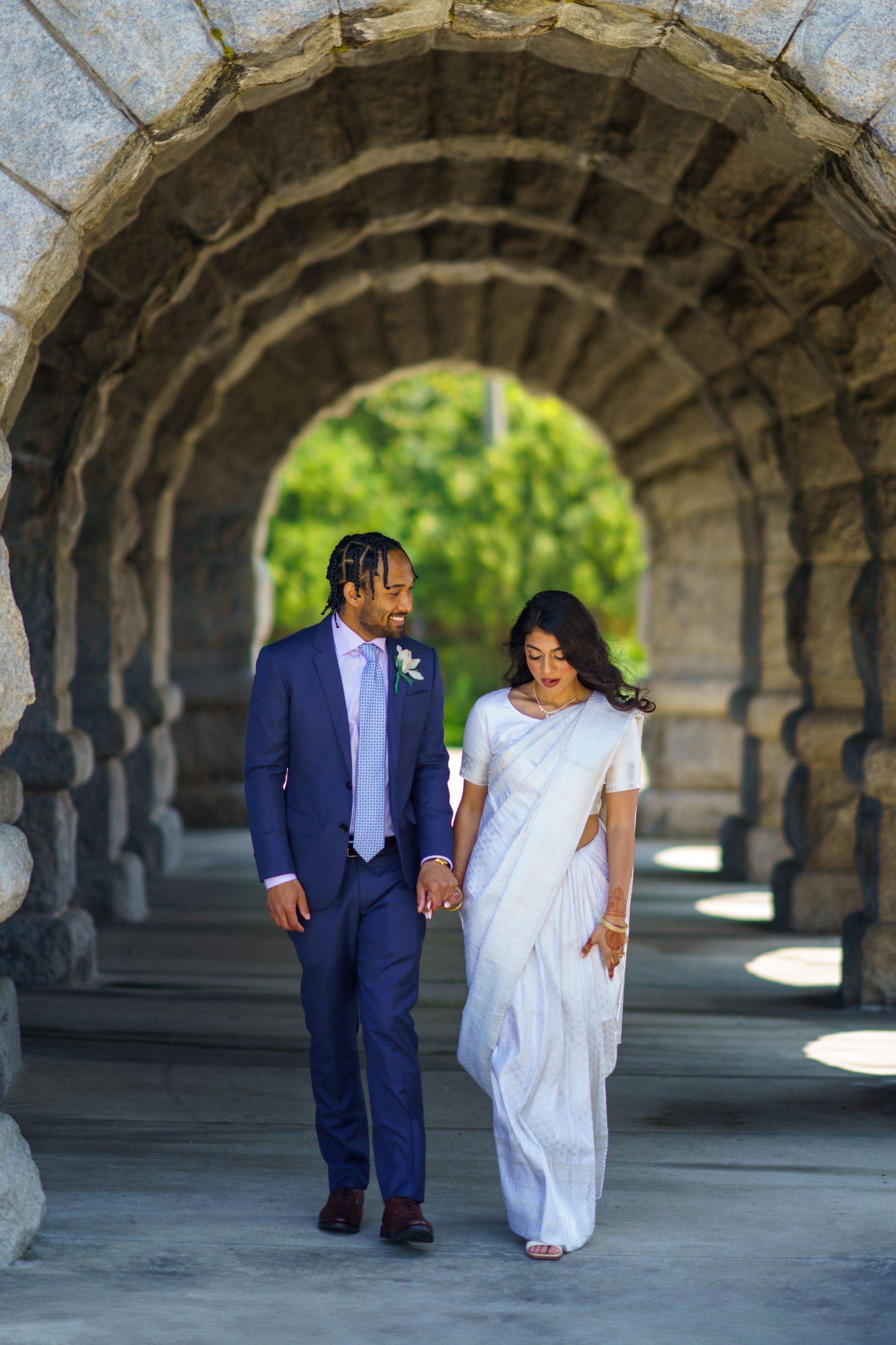 Couple in formal wear walking under a stone archway, holding hands, smiling.