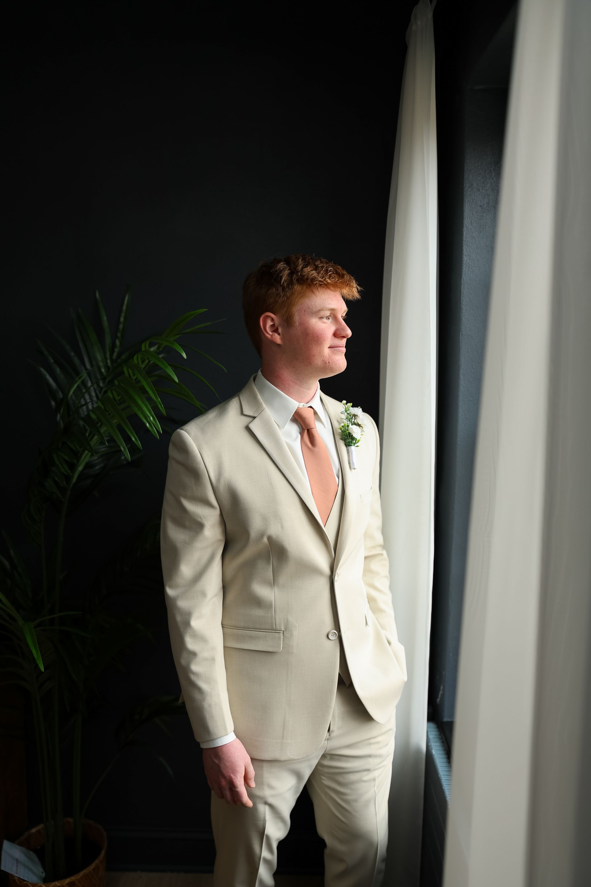 Man in a beige suit looks out a window. Dark wall and a plant on the left.