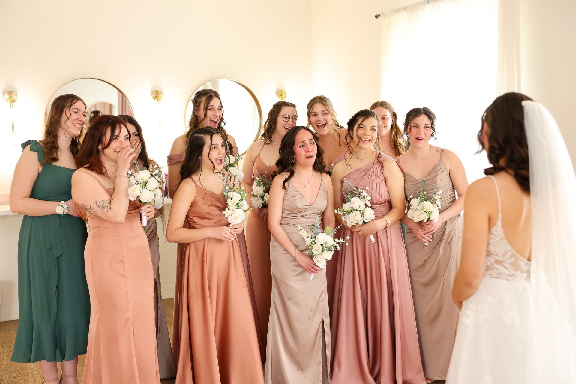 Bridesmaids react with delight as they see the bride in her wedding dress in a room with mirrors.