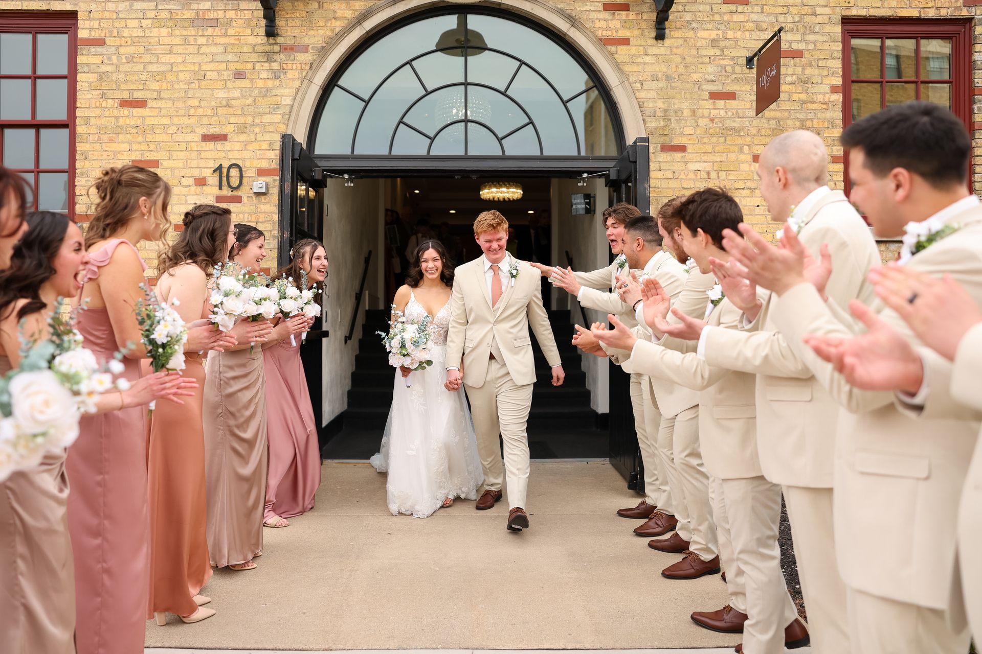 Newlyweds walk out of arched doorway, cheered on by wedding party. Building exterior, beige.