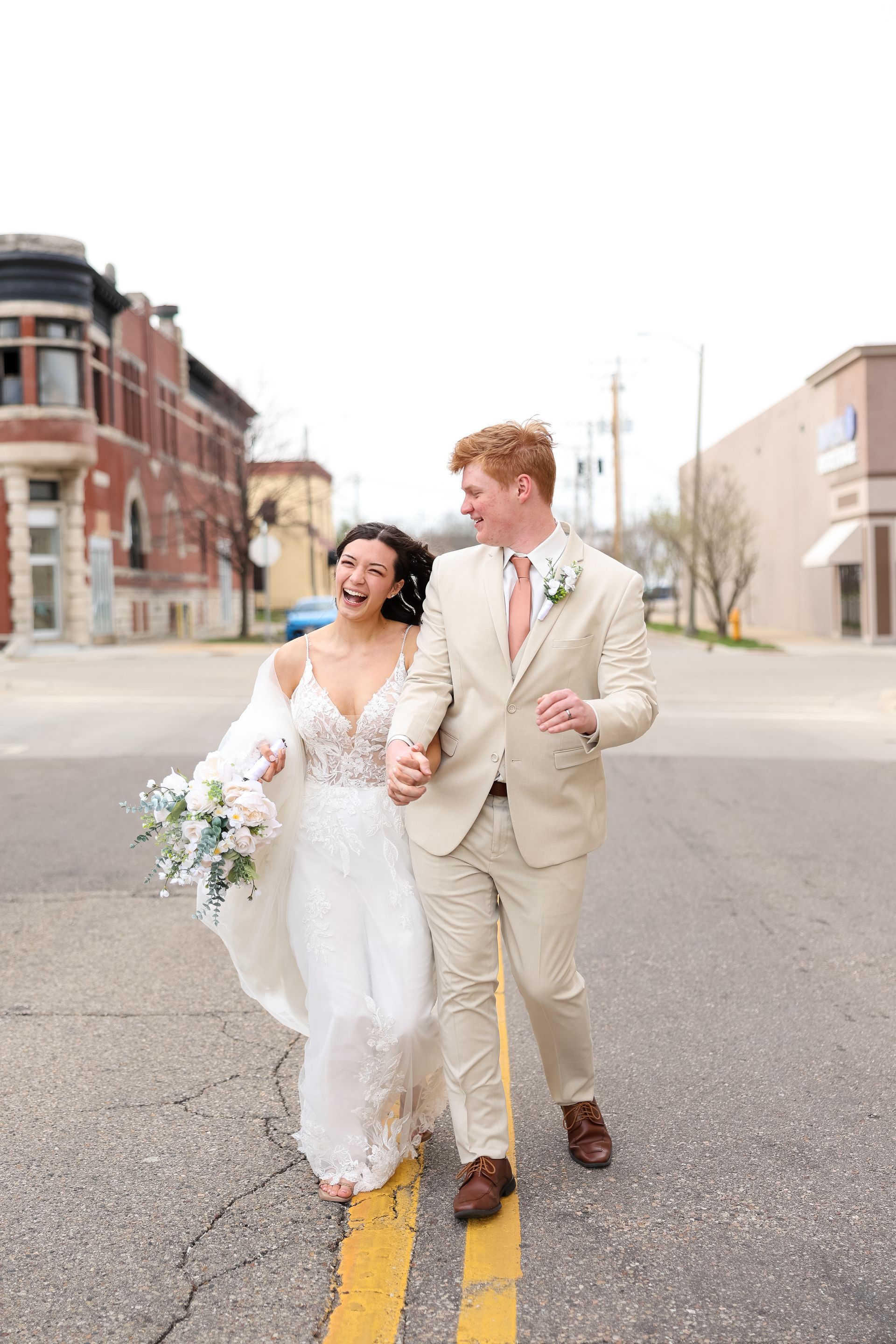 Newlyweds hold hands and walk down a street. The bride wears a white dress and the groom wears a tan suit.