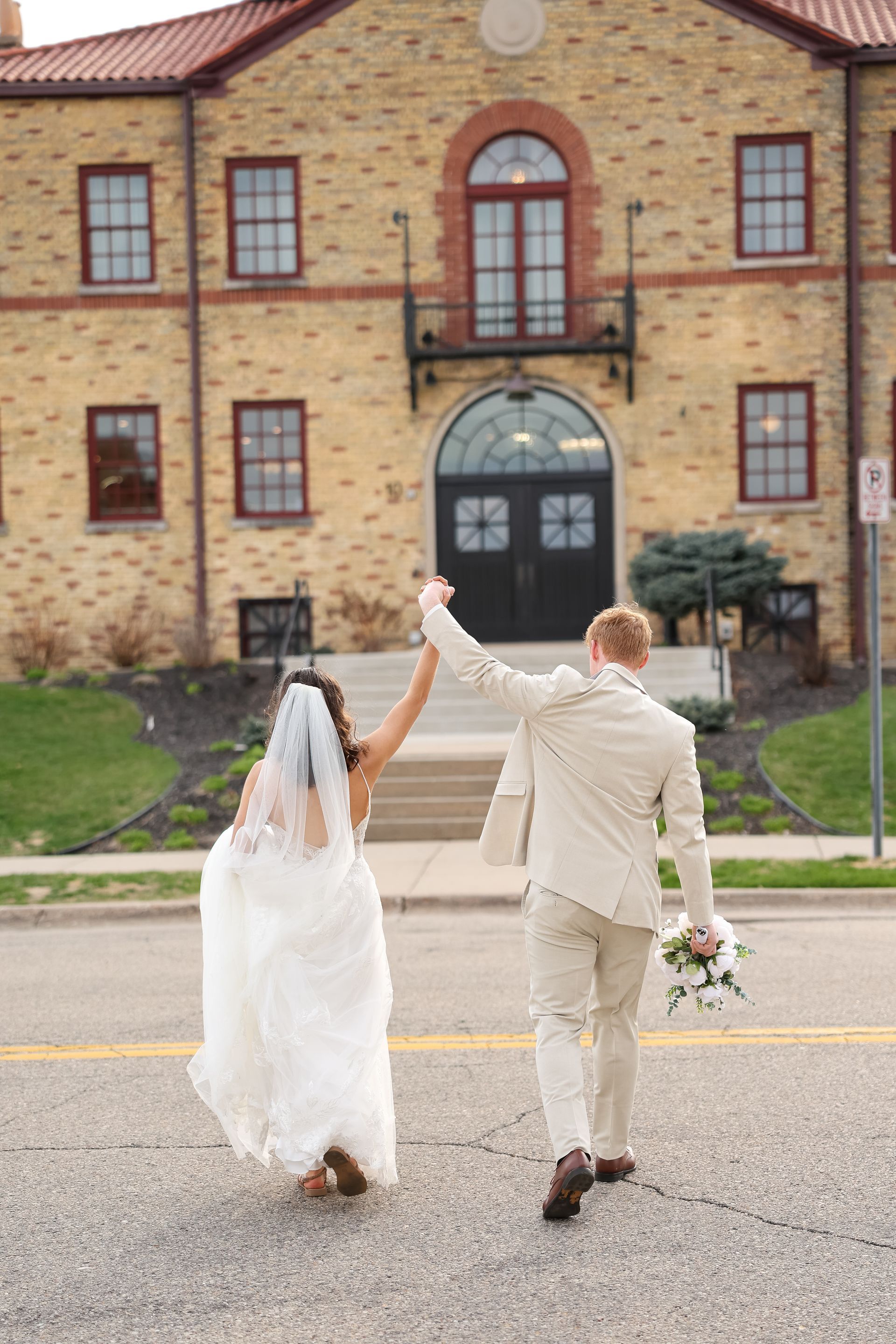 Bride and groom walking and holding hands in front of a brick building.