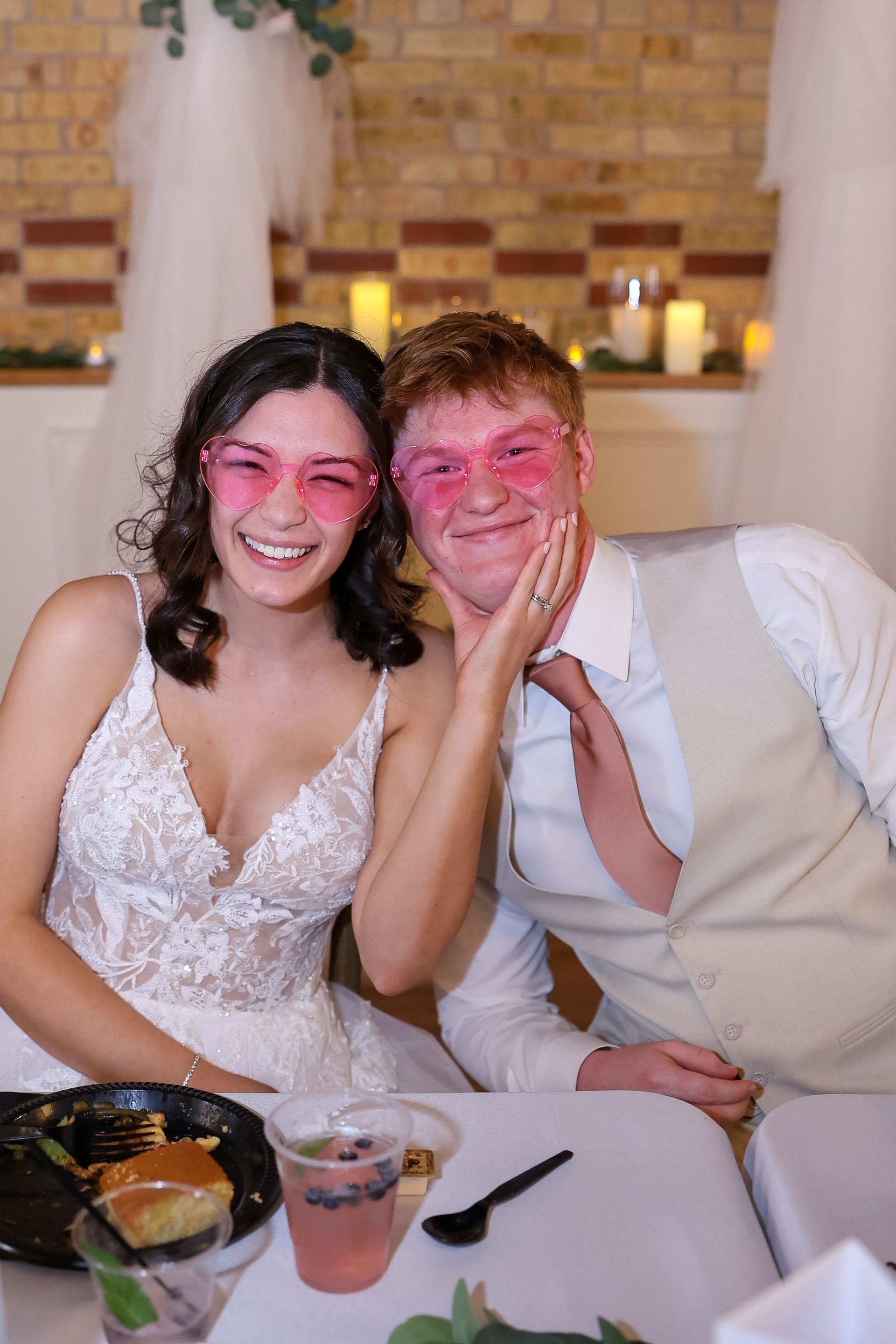Couple wearing pink heart-shaped glasses smiling, seated at a wedding reception table.
