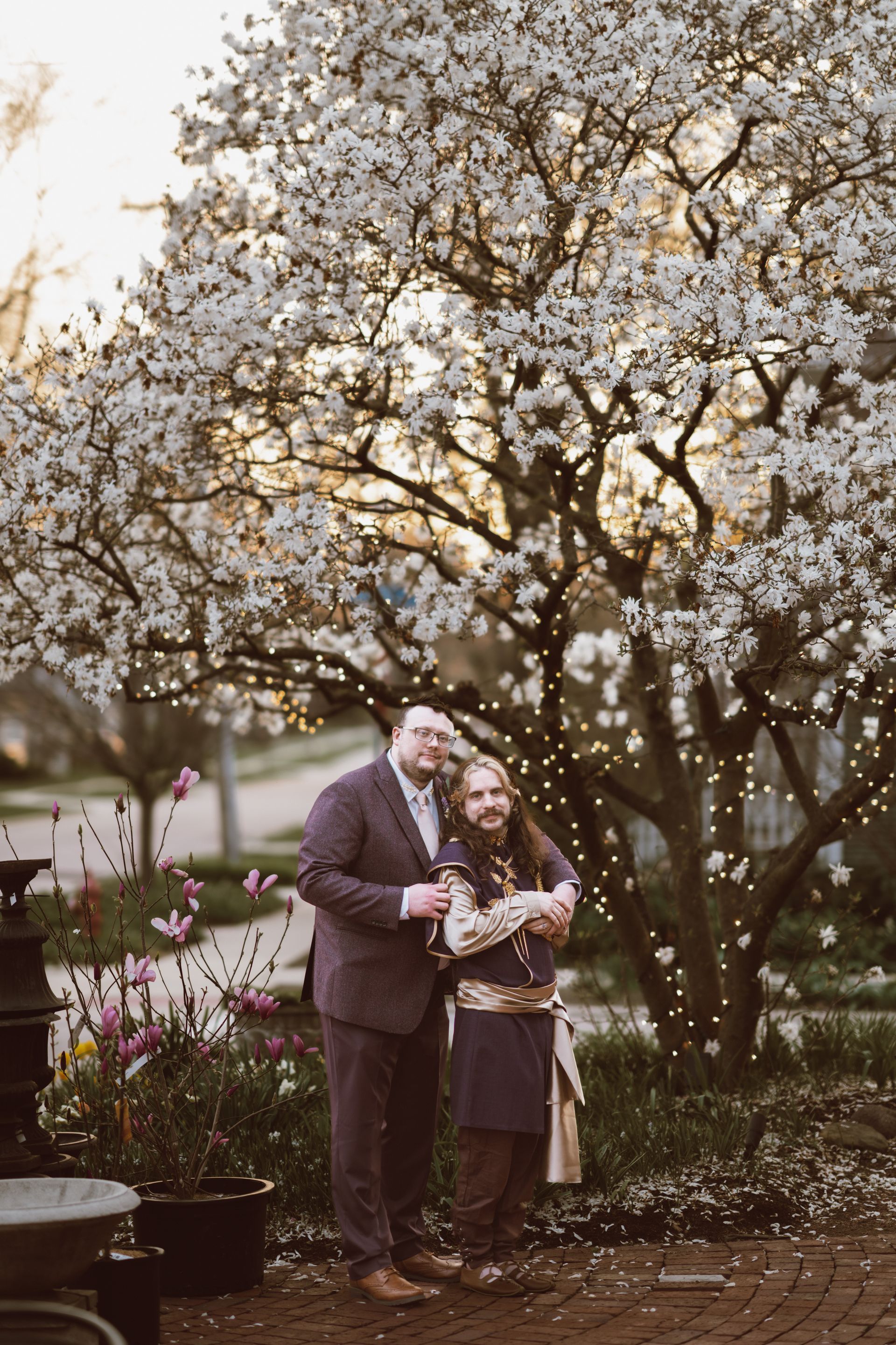 Two men embrace under a blooming tree. One in a suit, the other in earthy tones. Twinkling lights and floral backdrop.