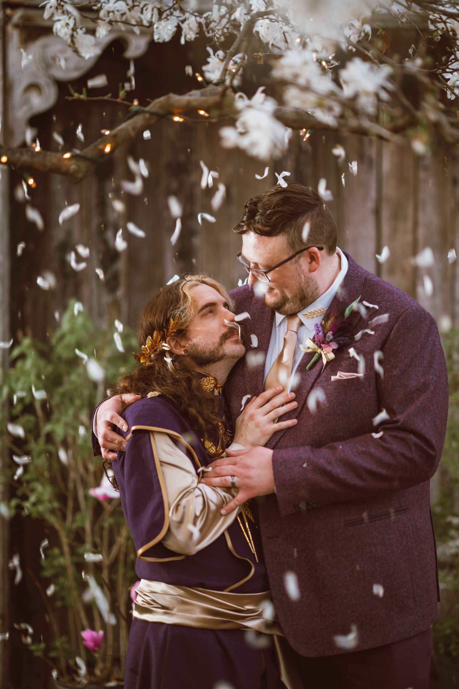 Two men embrace, confetti falling. One wears a purple tunic with floral hair, the other a purple suit, both smiling.