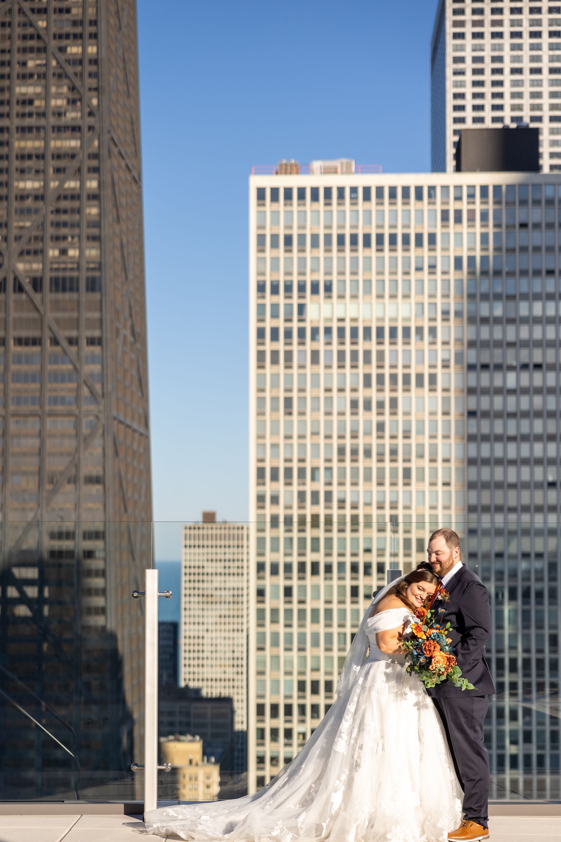 Bride and groom embrace on a rooftop in a city. Buildings and sky in background, fall colors in bouquet.