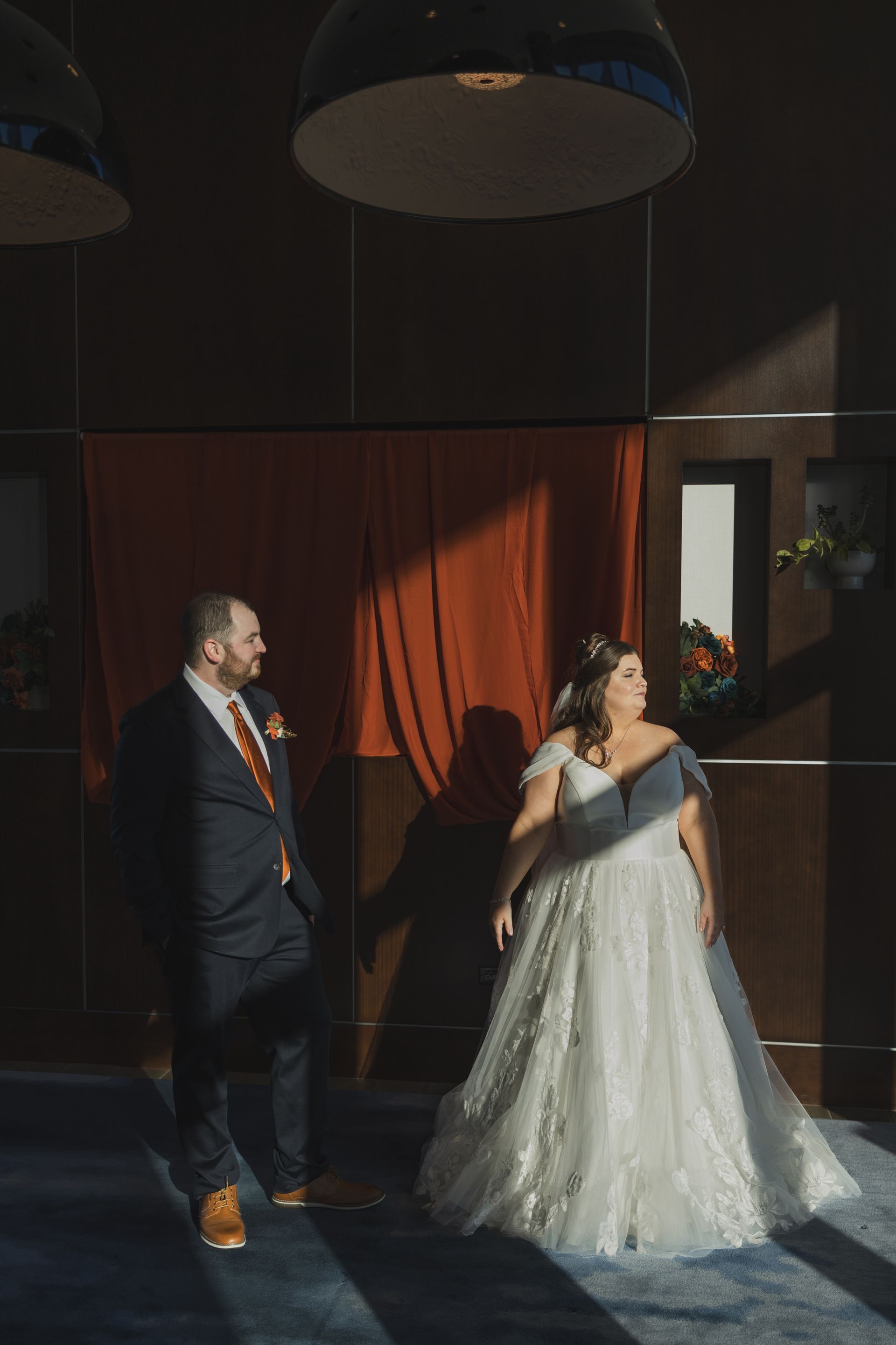 Bride in white dress gazes up, groom in navy suit looks on. Warm sunlight illuminates the couple and red curtain.