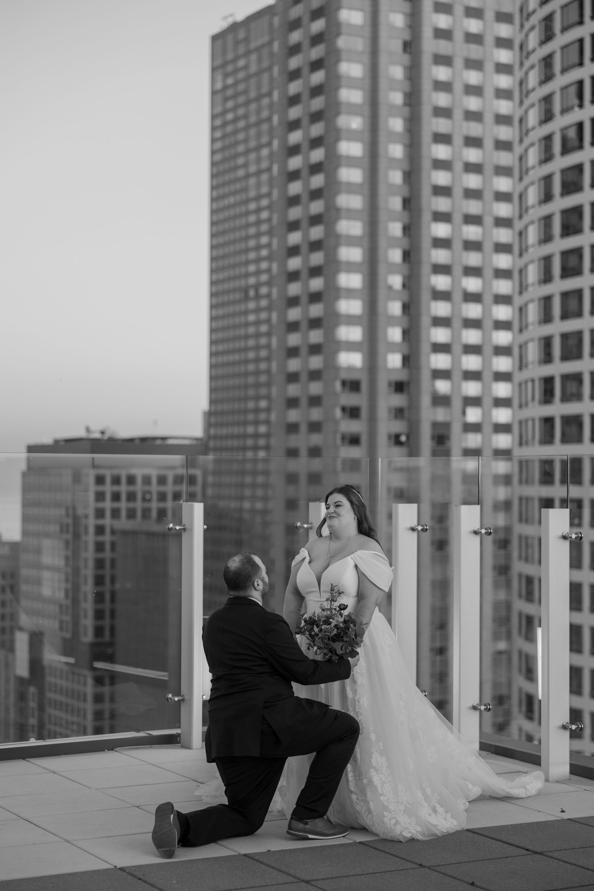 Man on one knee proposing to a person in a white dress on a rooftop with city buildings in the background.