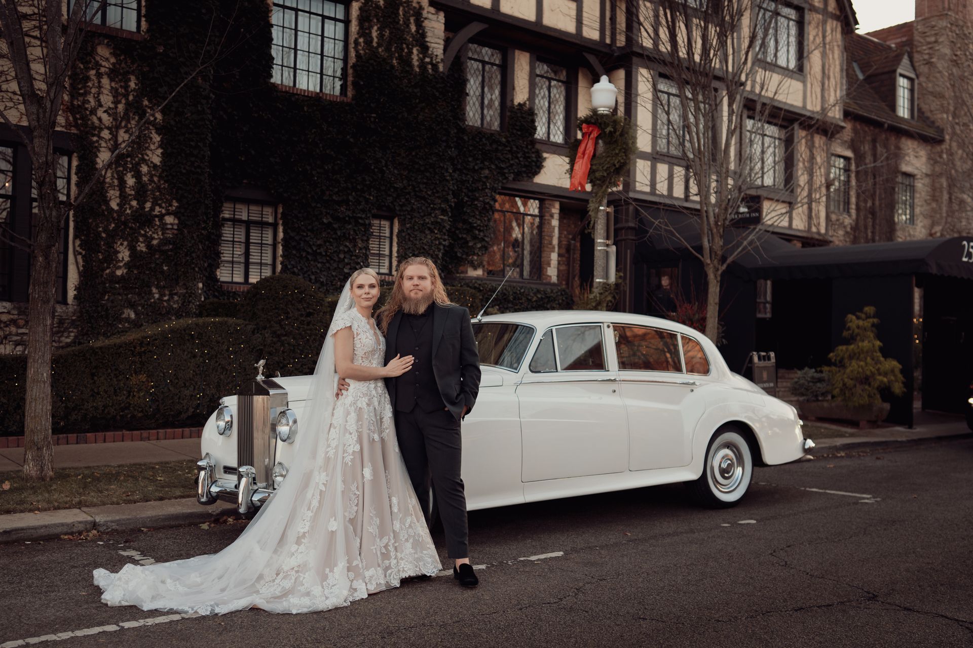 Bride and groom pose by a white vintage car in front of a Tudor-style building with ivy.
