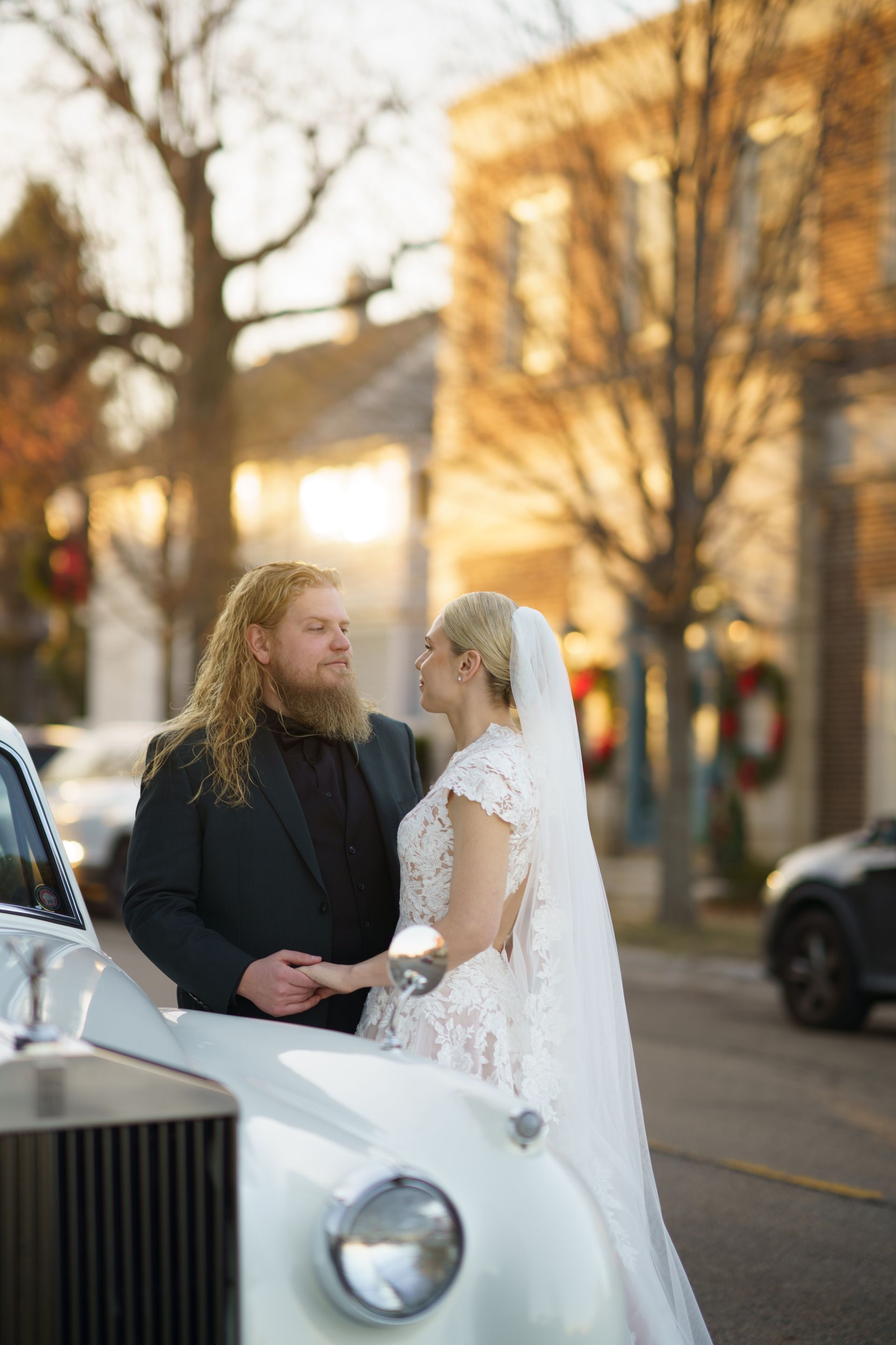 Bride and groom pose by a white vintage car on a street. The couple looks at each other, bathed in sunlight.