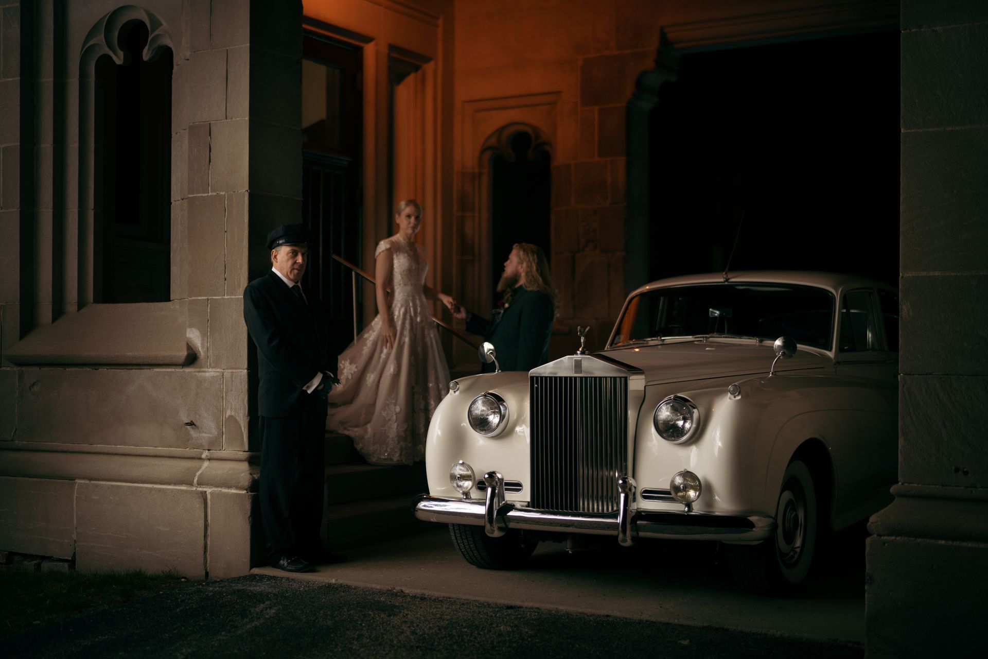 White Rolls Royce car parked in front of a stone building. A woman in a gown exits the car. A man in uniform waits.